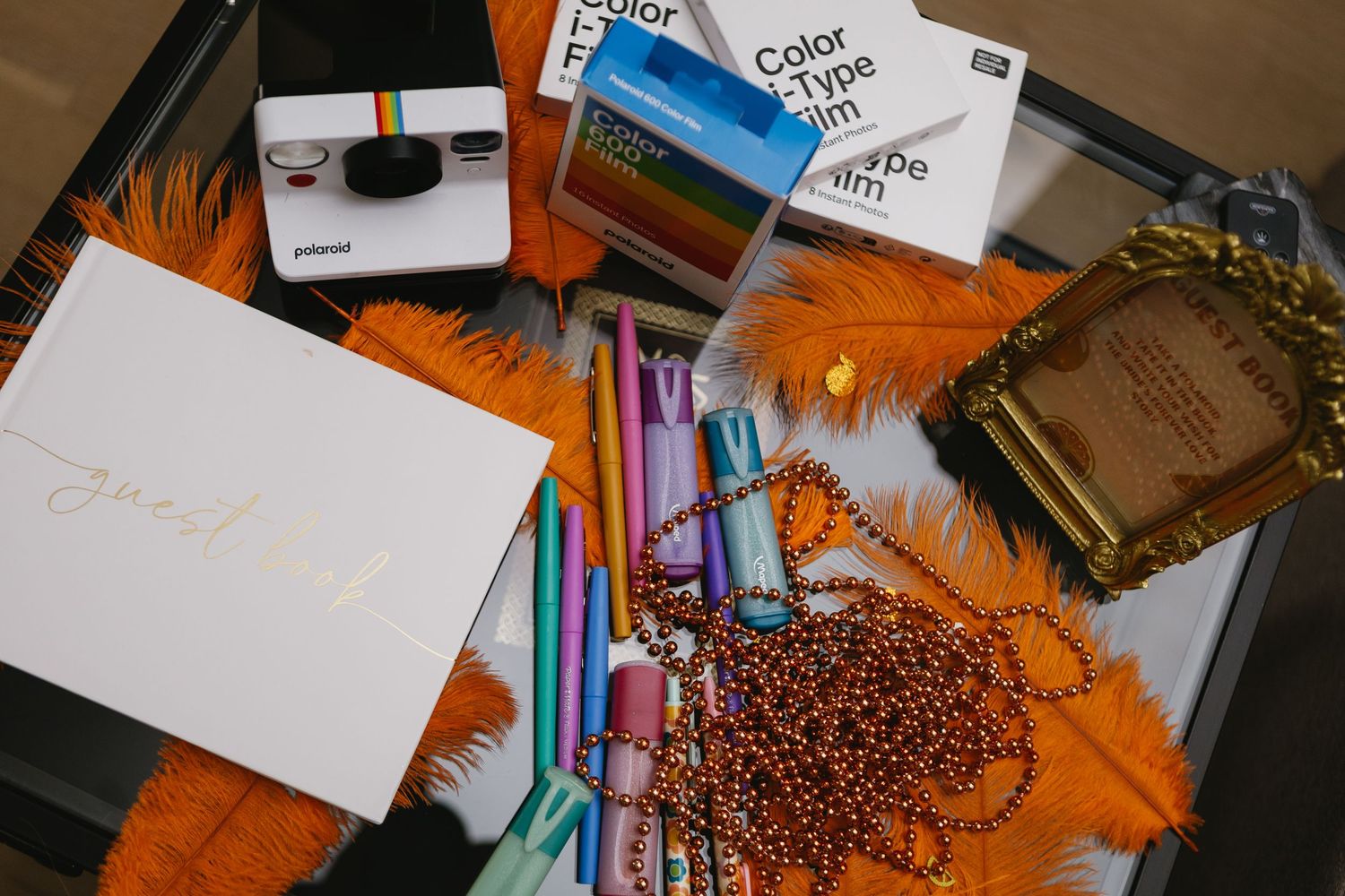 Flat lay of Polaroid camera, color film, guest book, pens, orange feathers, and gold frame on dark tray.