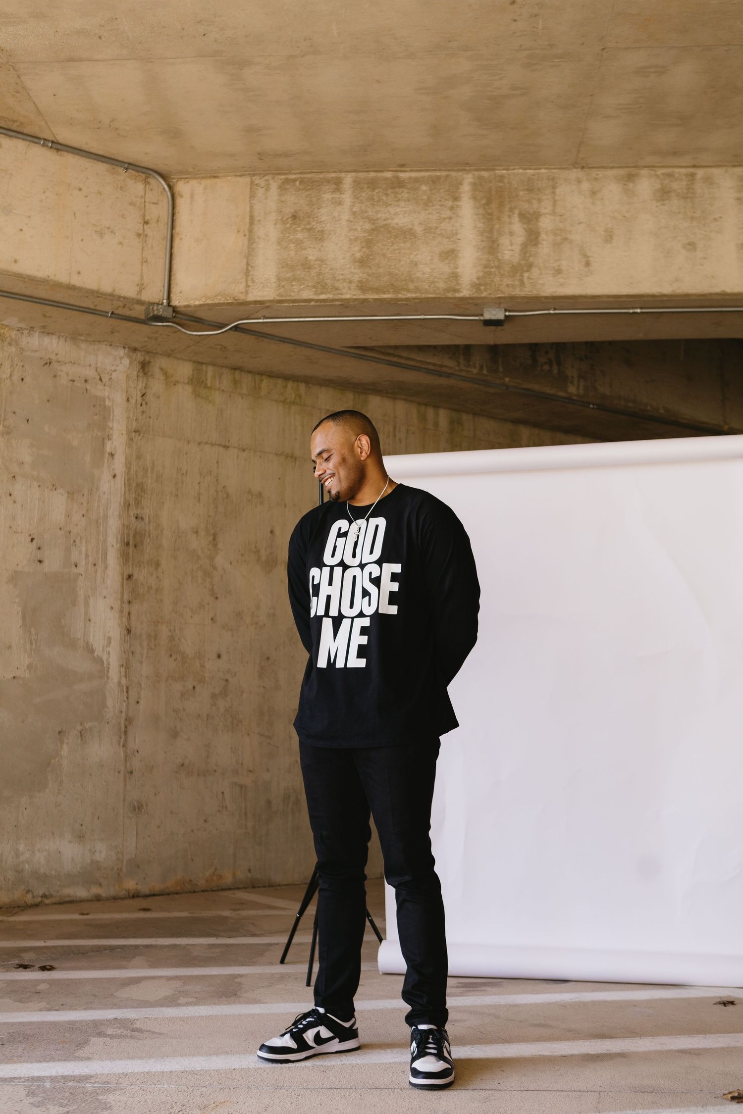 Man in black GOD CHOSE ME sweatshirt and pants smiling, standing in a concrete parking structure.
