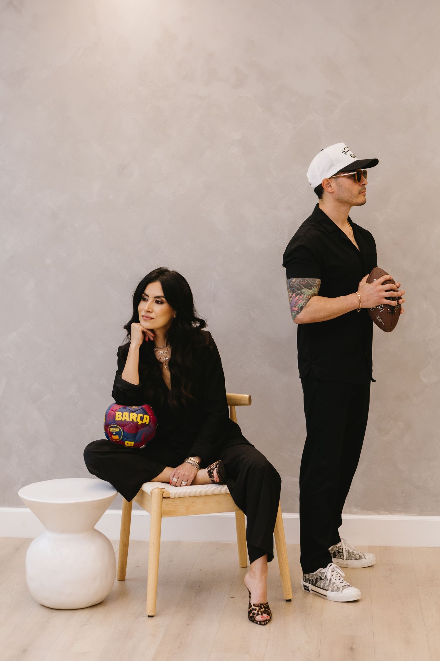 A woman in black sits on a wooden chair holding a Barca bag while a tattooed man in black stands holding a football.