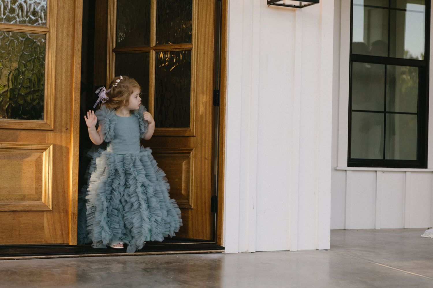 Young girl in a ruffled teal flower girl dress standing at a wooden door entrance of a white building.