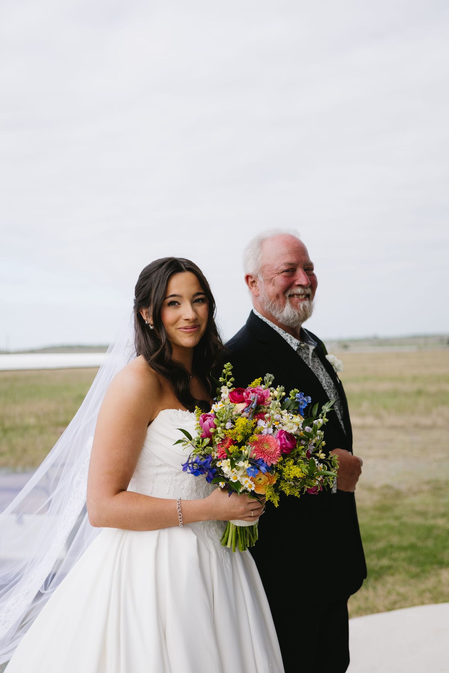 Bride in white strapless gown holding colorful bouquet stands with older gentleman in black suit outdoors.