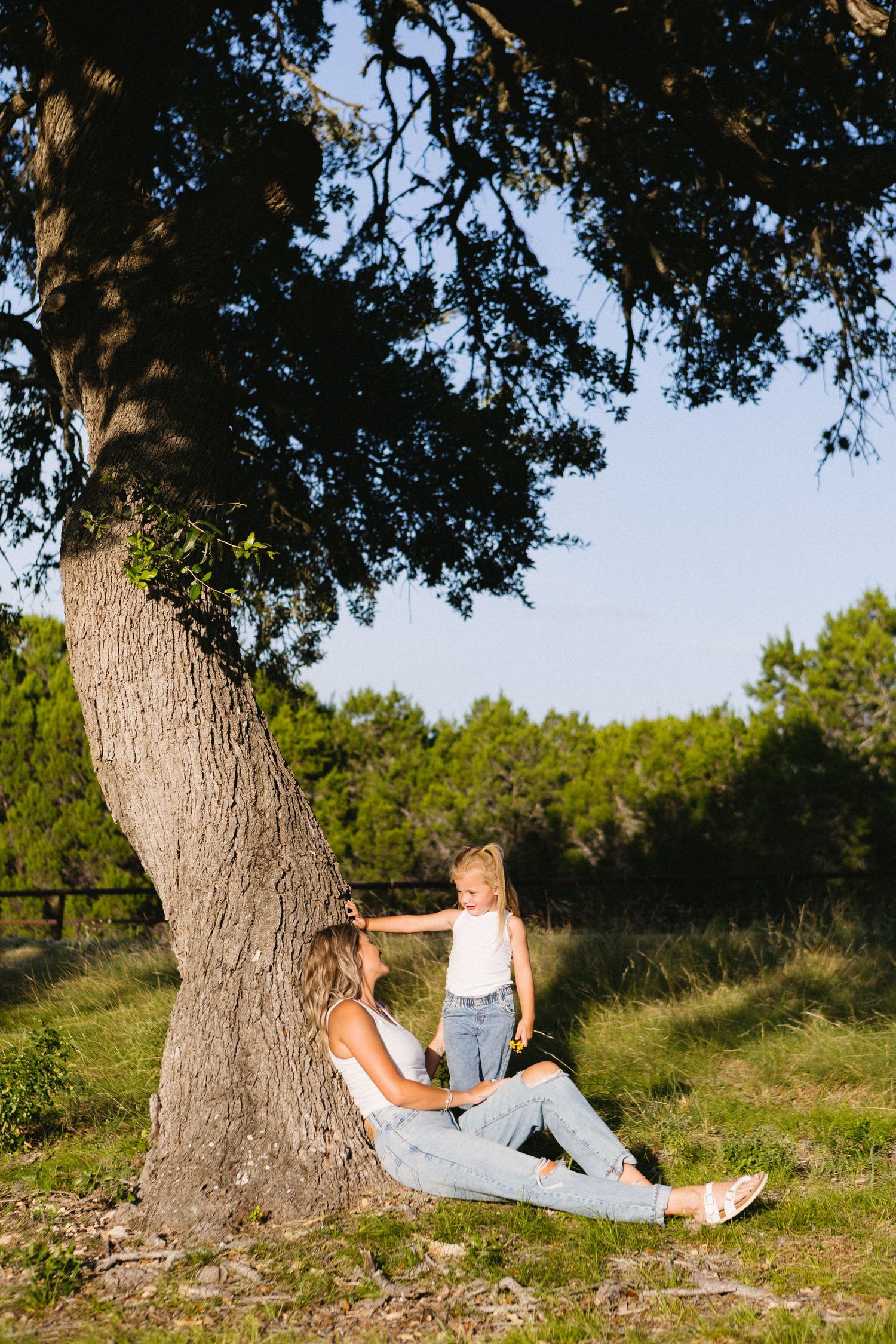 A mother and young daughter share a tender moment sitting under a large oak tree in a sunny green meadow.
