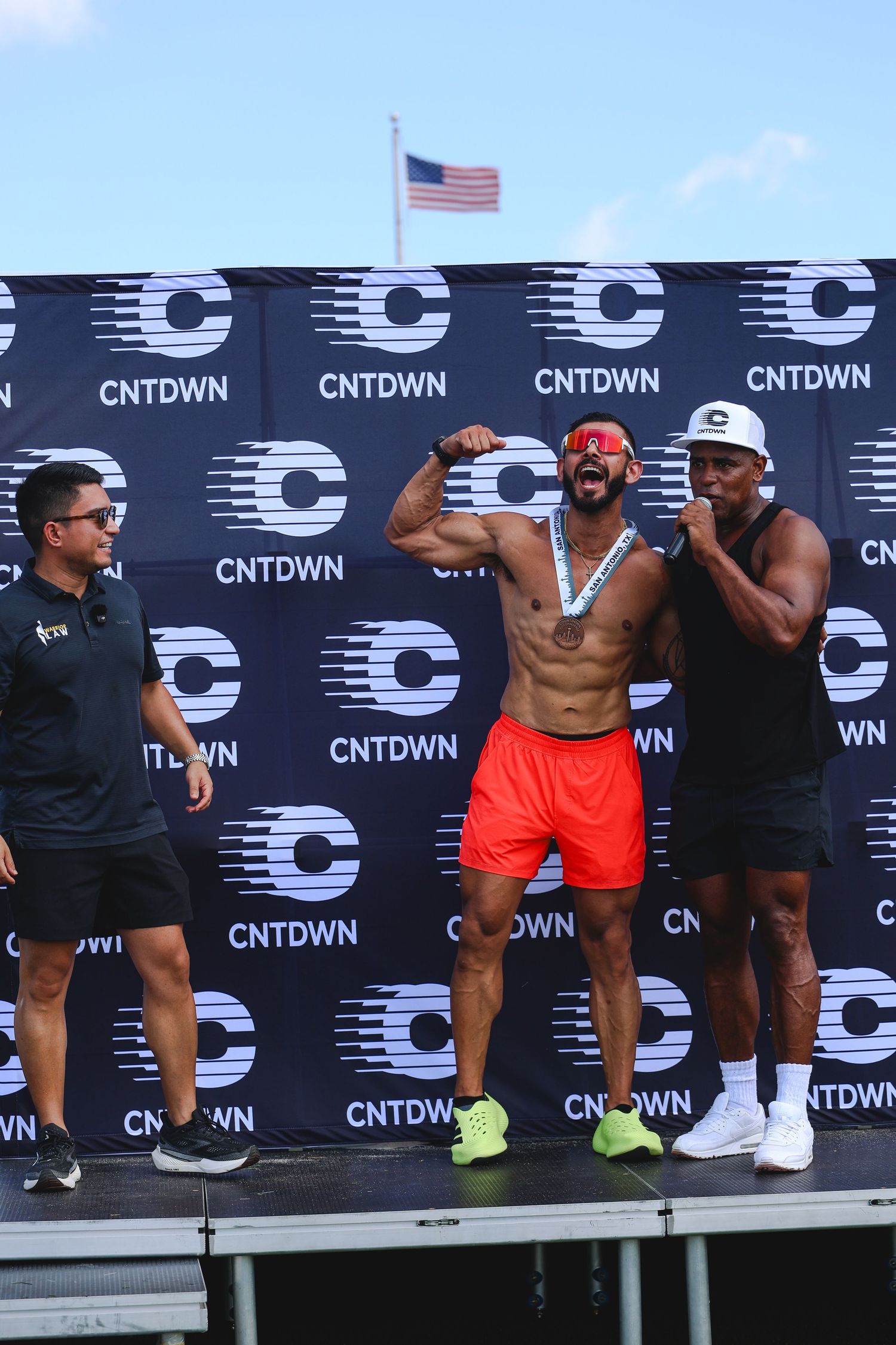 Shirtless athlete flexes on CNTDWN event stage wearing medal and red shorts, flanked by two men.