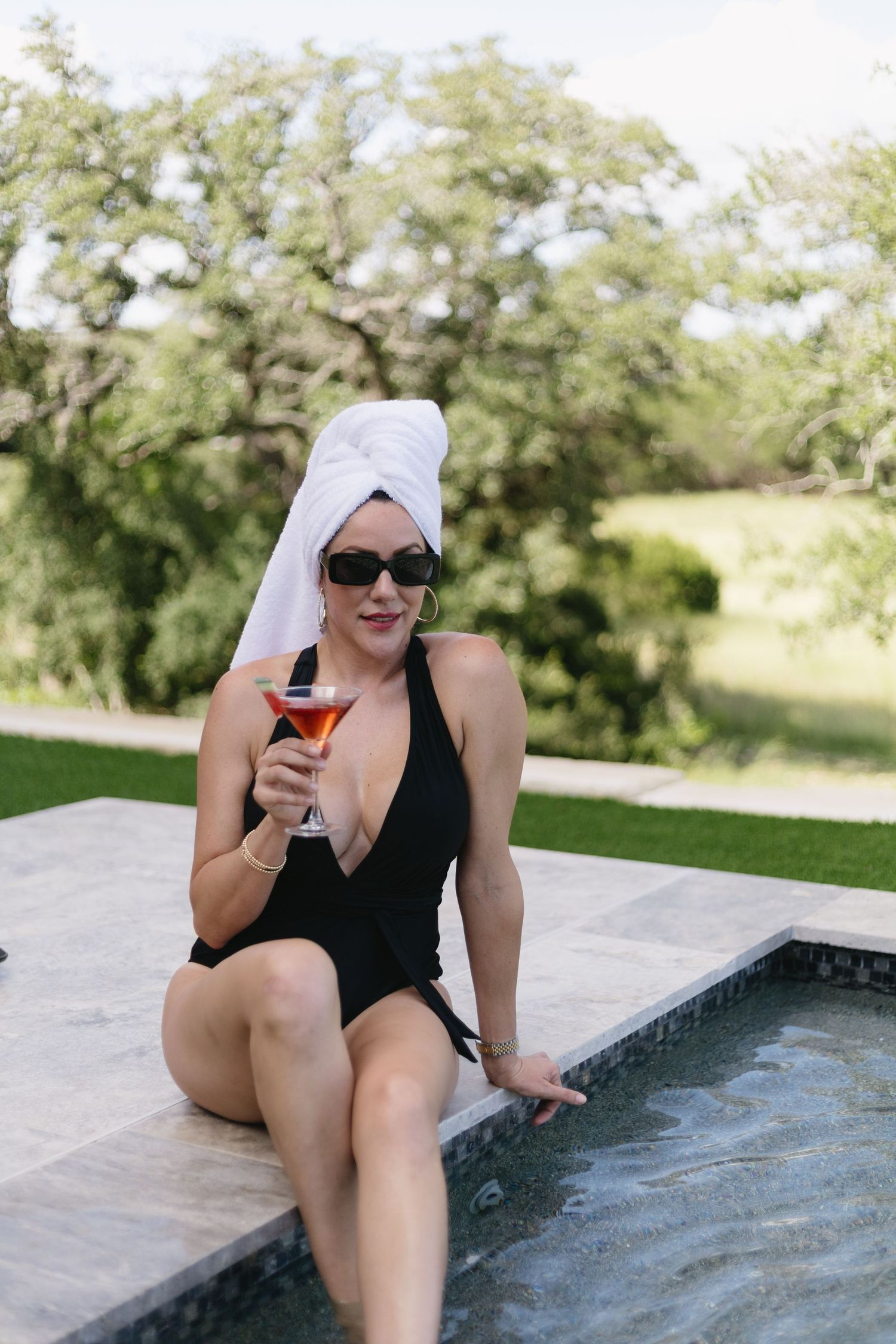 Someone lounges by a modern pool edge wearing a black swimsuit and white headwrap while holding a drink on a sunny day.