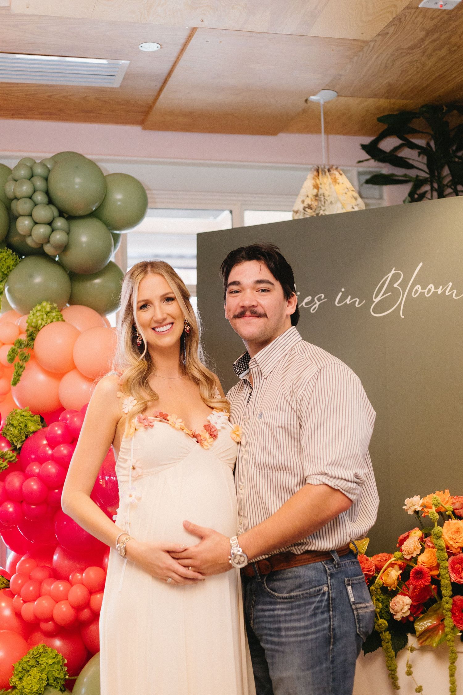Couple poses at a baby shower with colorful balloon arch and floral decorations.