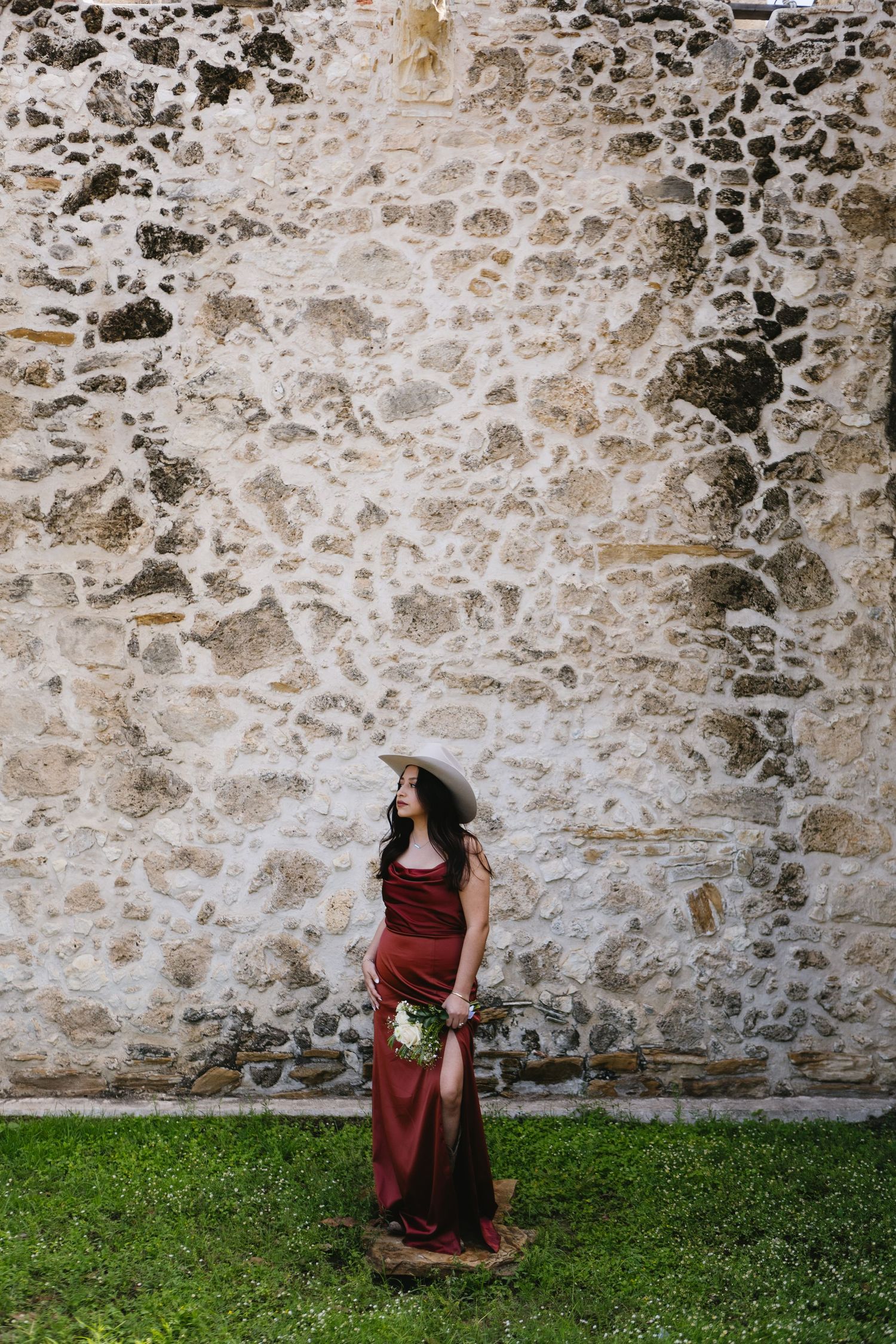 Woman in red slit dress and wide-brim hat holding bouquet stands against rustic stone wall on green grass.