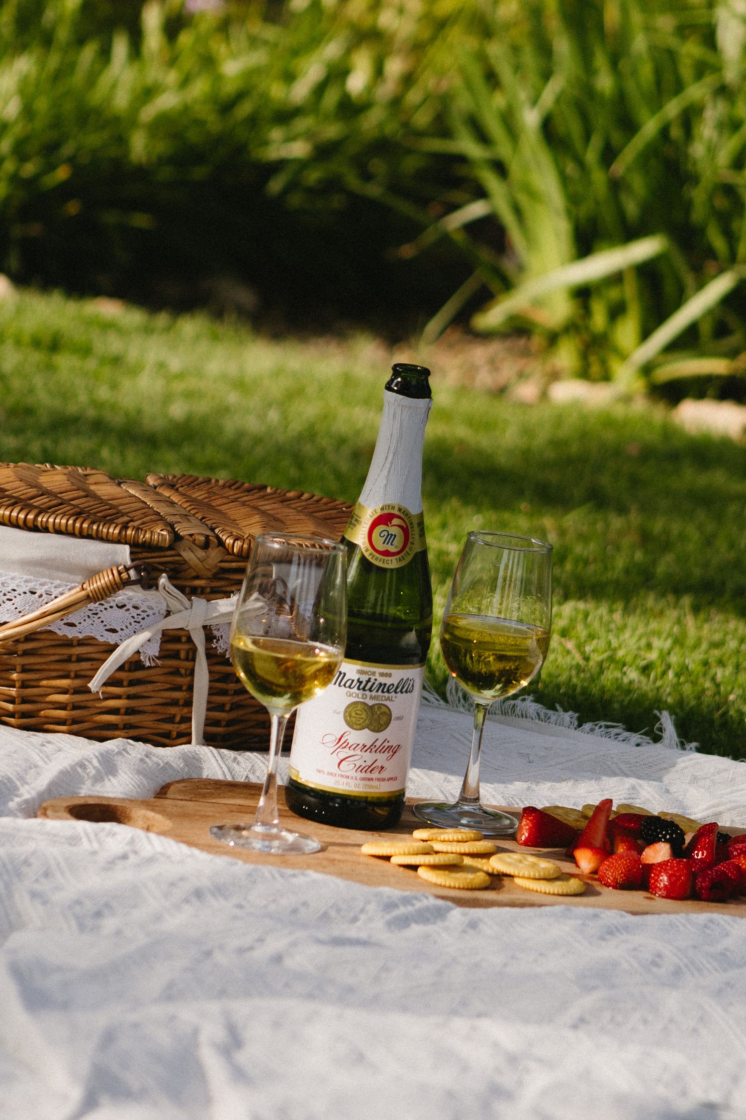 Martinelli's sparkling cider bottle with two glasses, strawberries, crackers on a wooden board at a garden picnic.