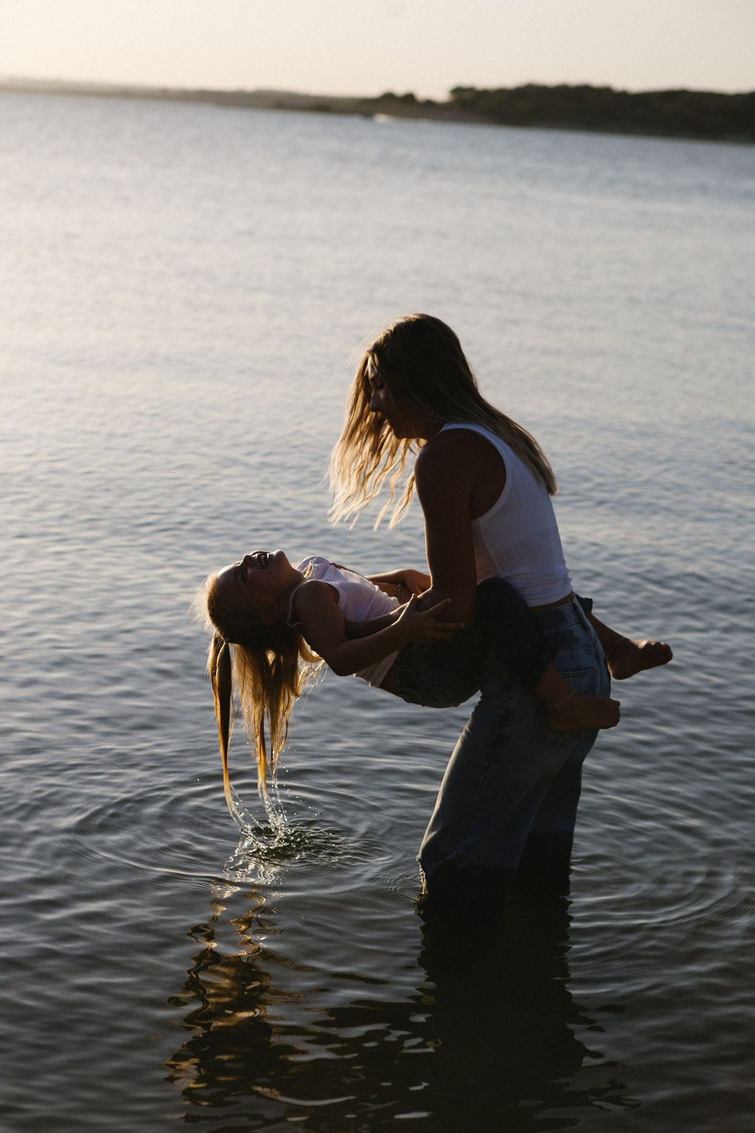 Two women playing in calm lake waters at sunset, one lifting the other in a joyful embrace.