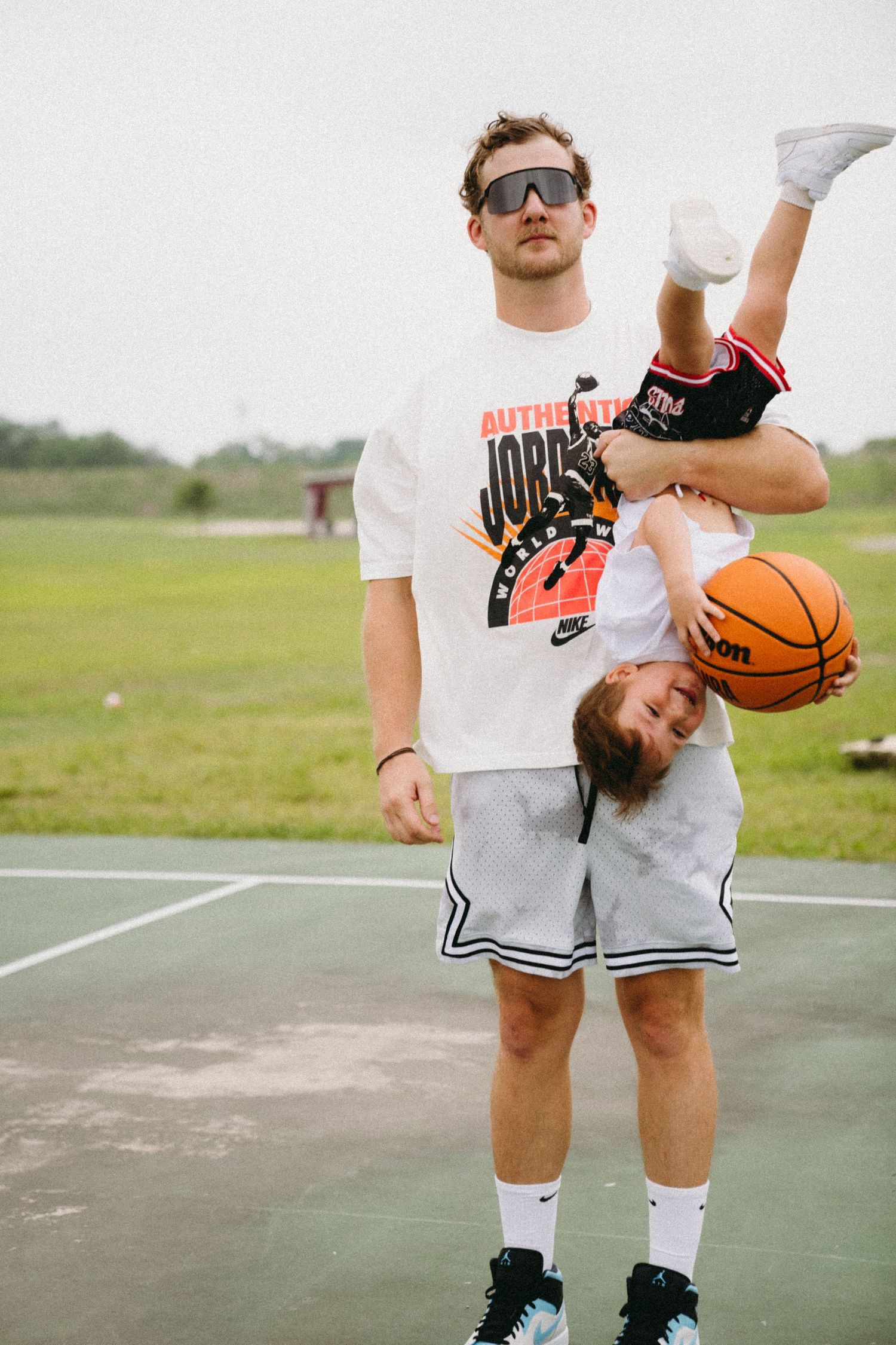 Man in Jordan shirt holds child upside down while child grips basketball on outdoor court.