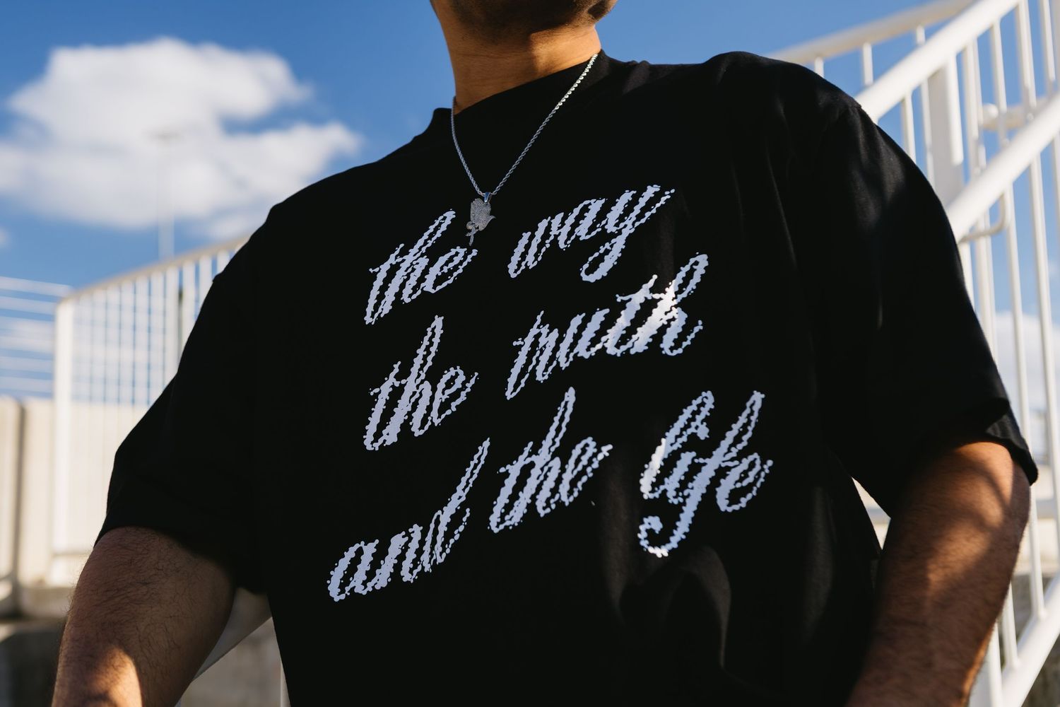 Man wearing black graphic tee with 'the way the truth and the life' script lettering and silver chain necklace.