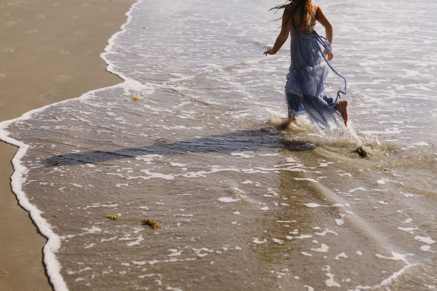 Woman in blue dress walking along ocean shoreline with waves washing over her feet on sandy beach.