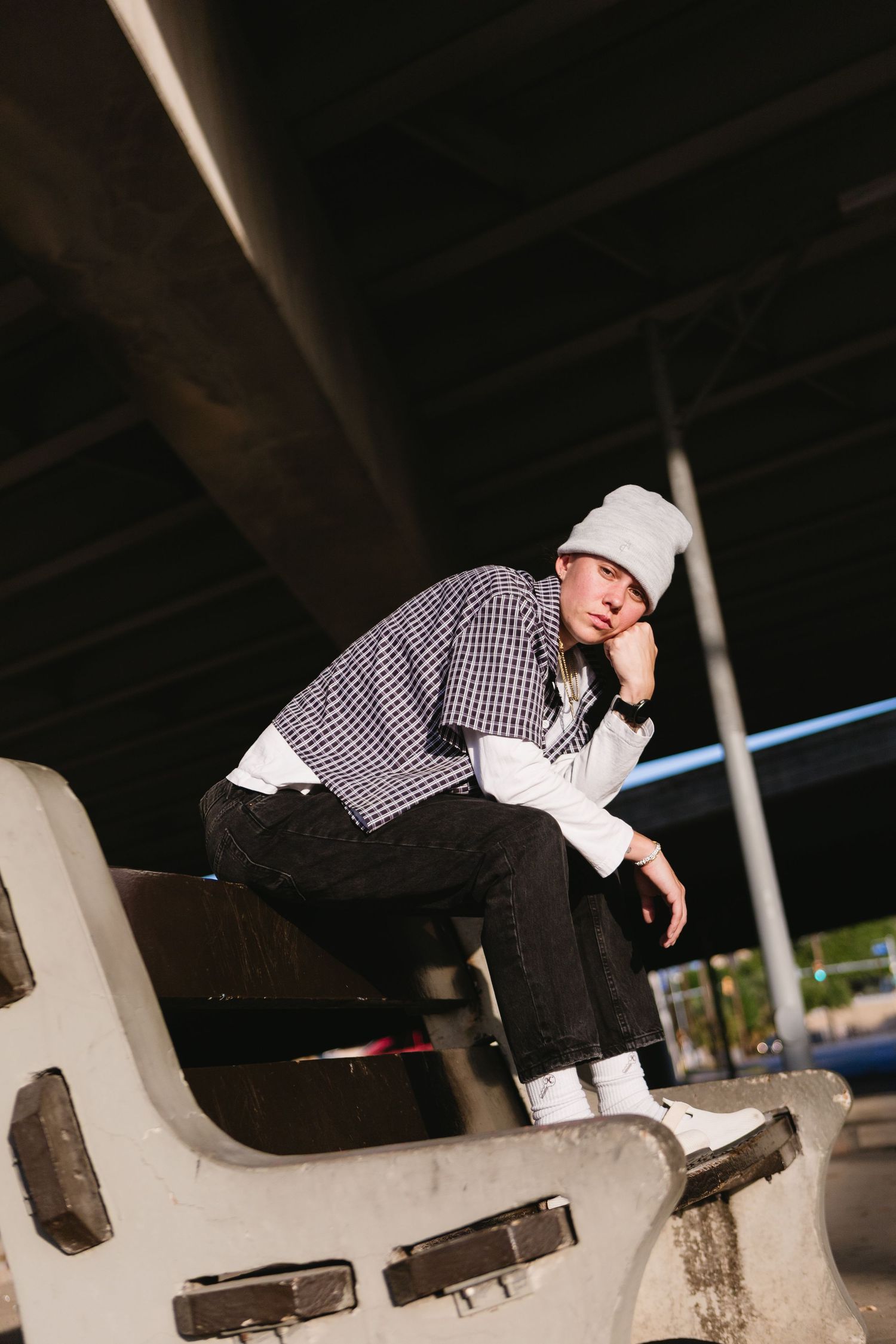 Young man in plaid shirt, black jeans, and gray bucket hat sitting on concrete barrier under bridge.