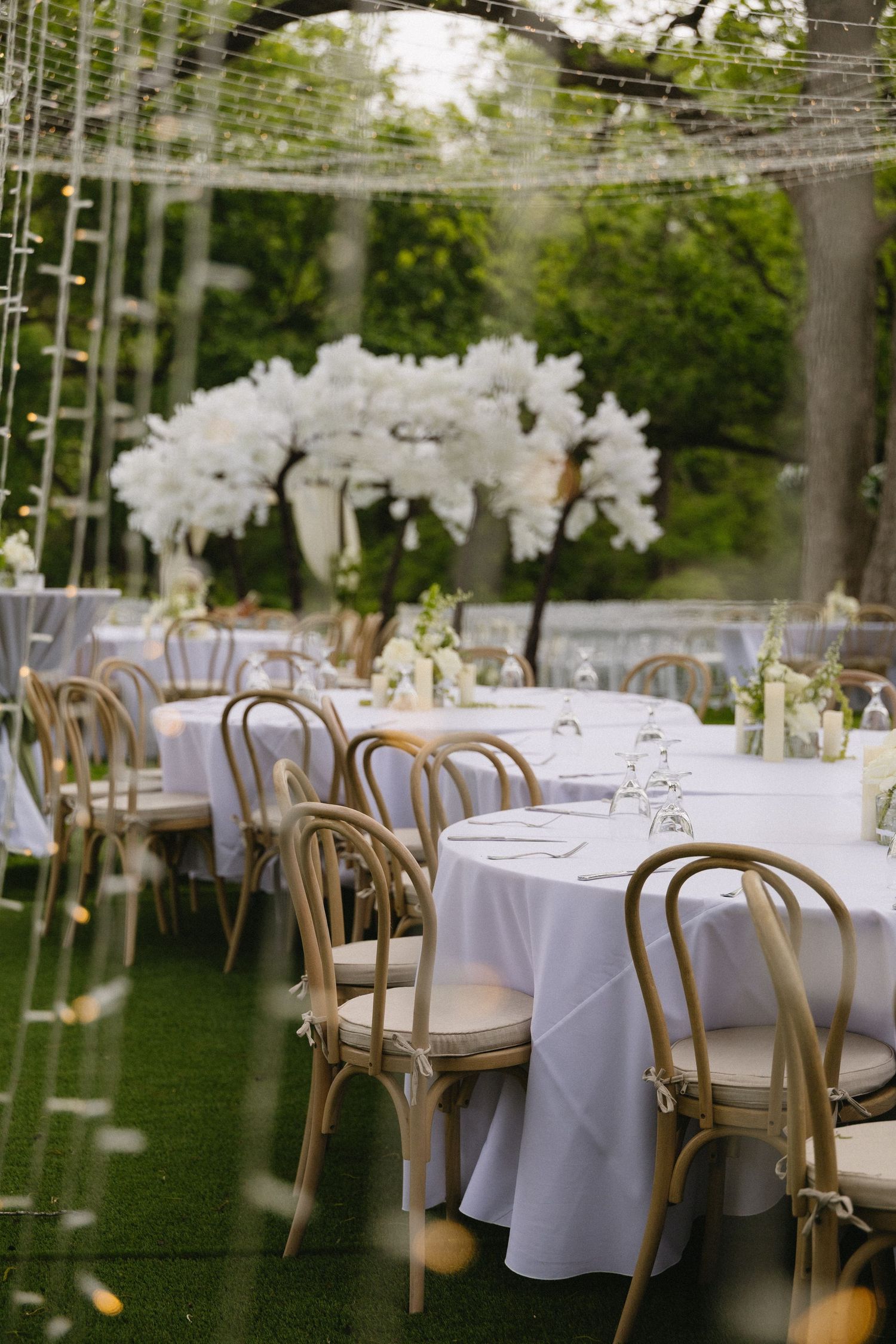 Elegant outdoor wedding reception with white linen tables, wooden cross-back chairs, and white floral tree centerpiece on green lawn.