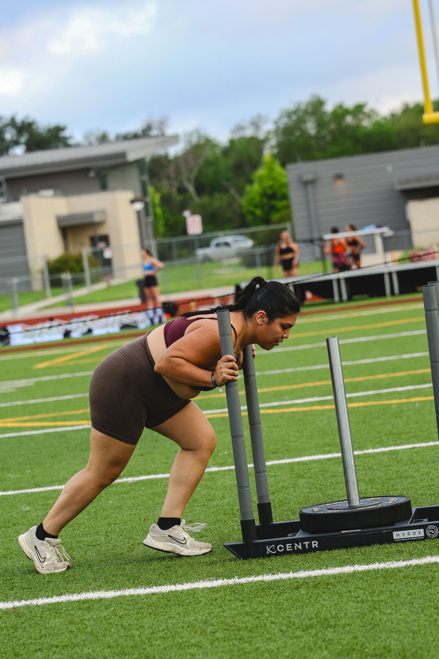 Woman in brown athletic wear pushes a KCENTR weighted sled on a green turf field during an outdoor workout.