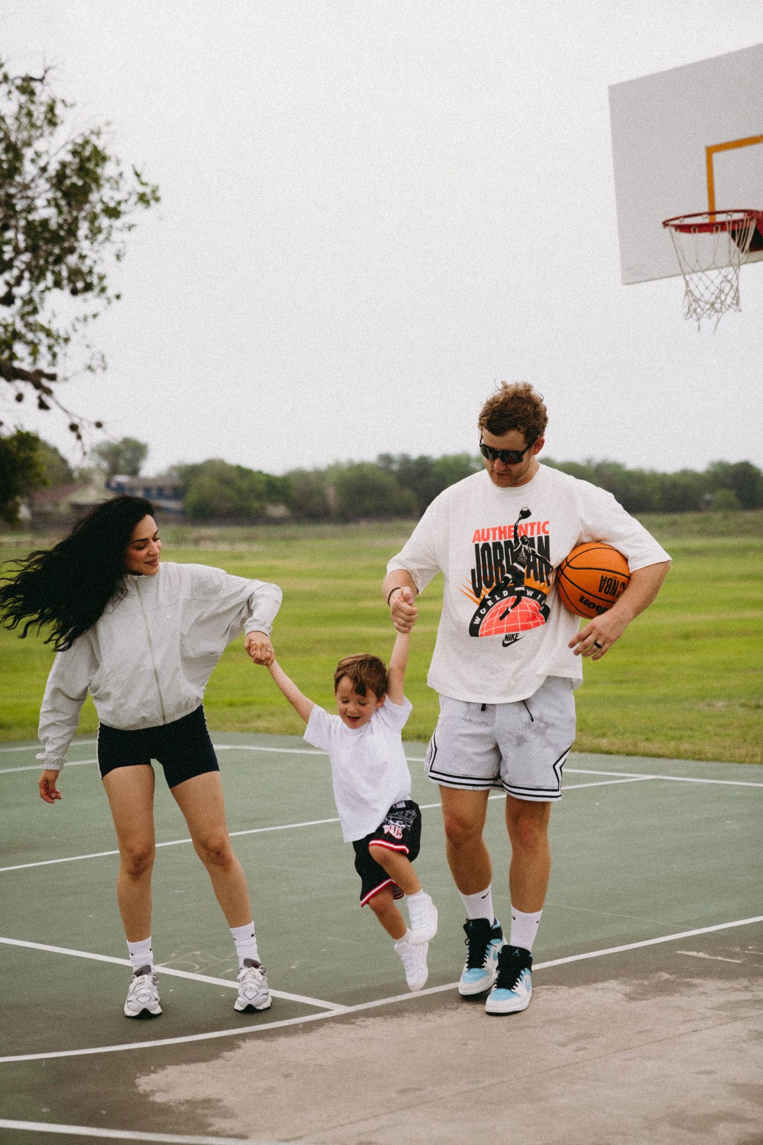 Family playing basketball outdoors, parents swinging child between them near hoop on court.