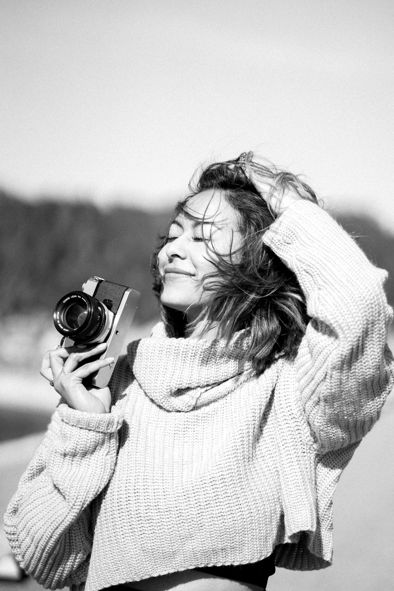 Black and white photo of a young woman in a cozy knit sweater holding a vintage camera outdoors on a windy day.