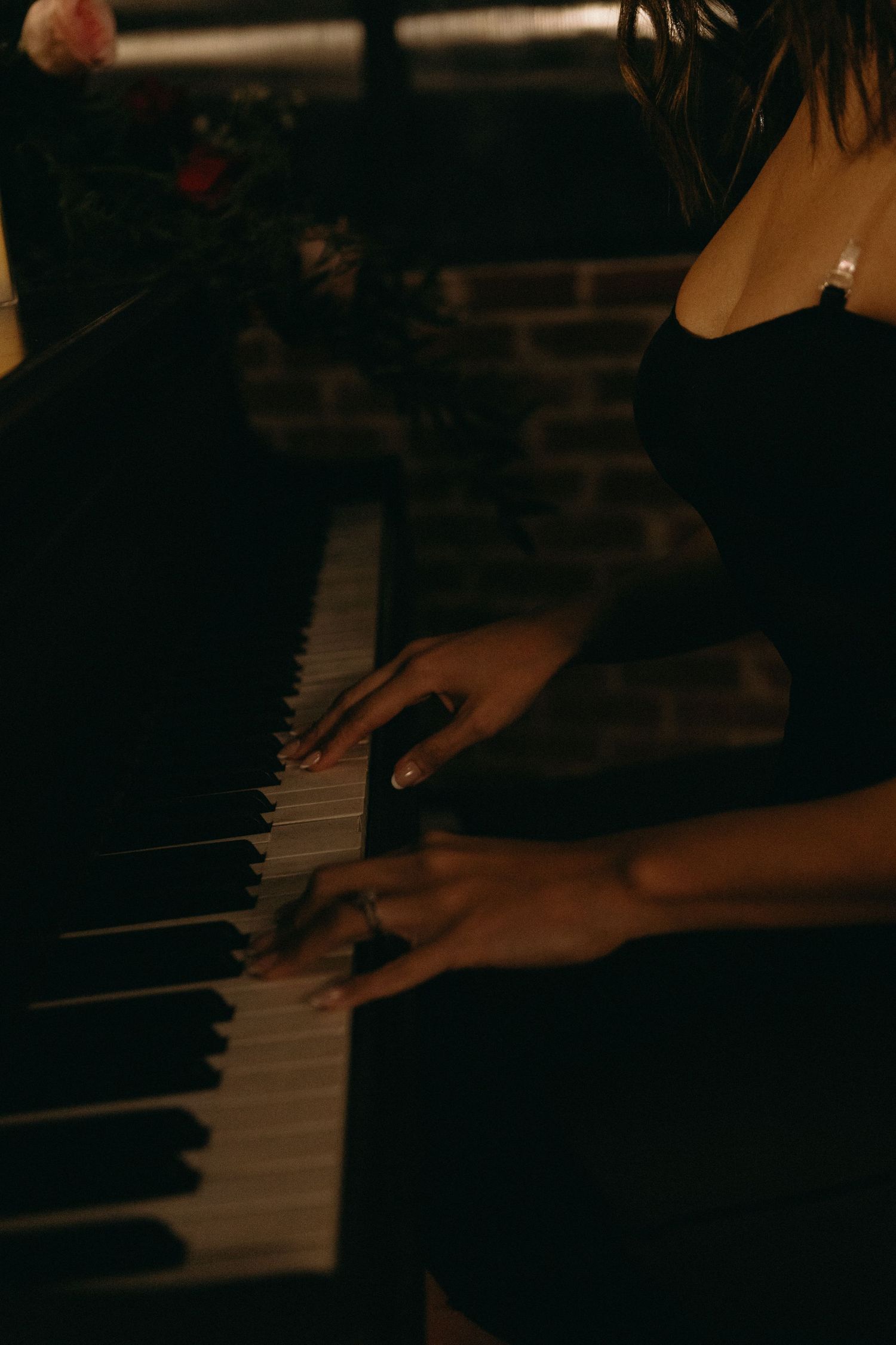 Woman in black dress playing piano keys in dim candlelit room, wearing rings, with flowers in background.