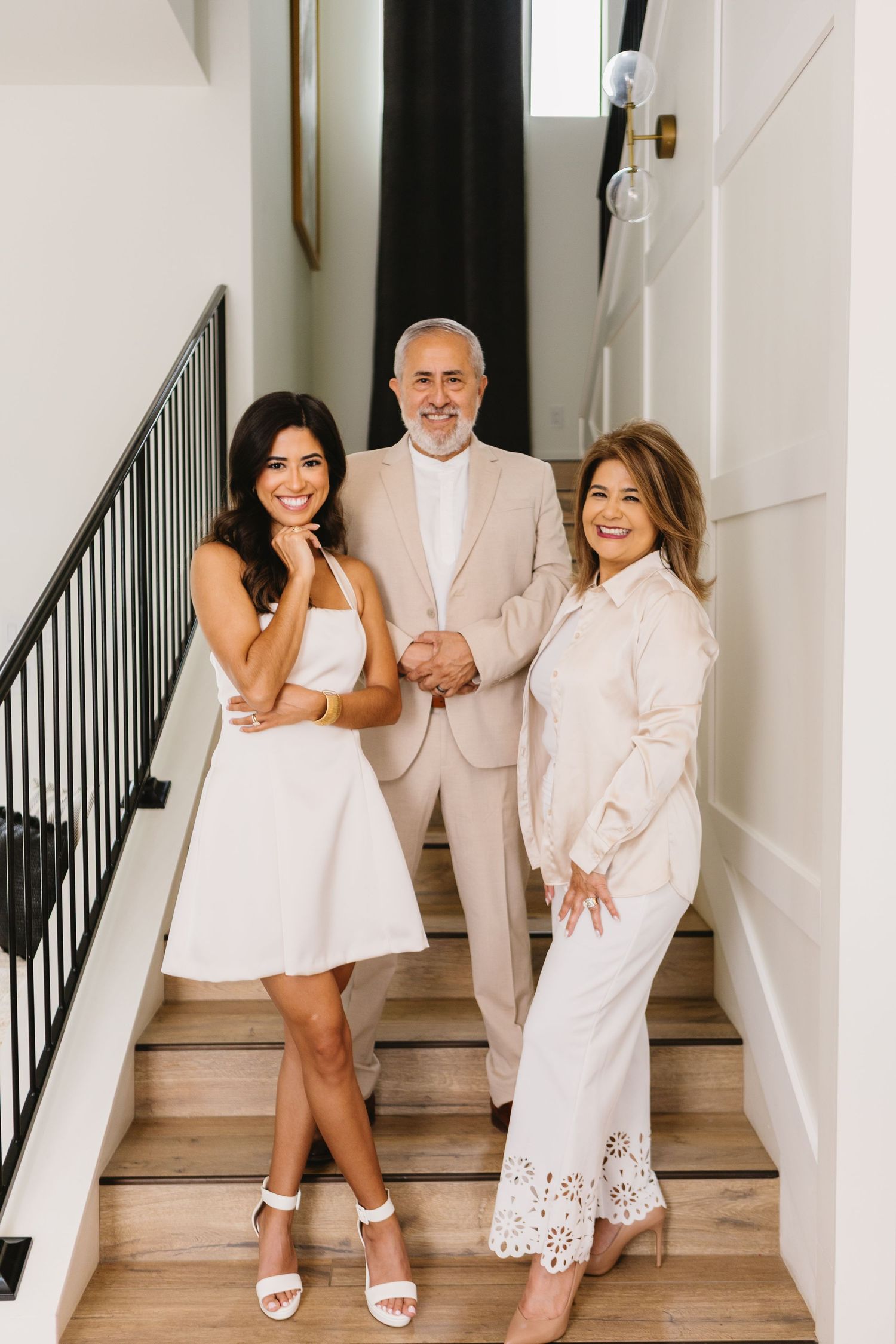 Three elegantly dressed people in white and beige outfits posing on a modern staircase with black railings.