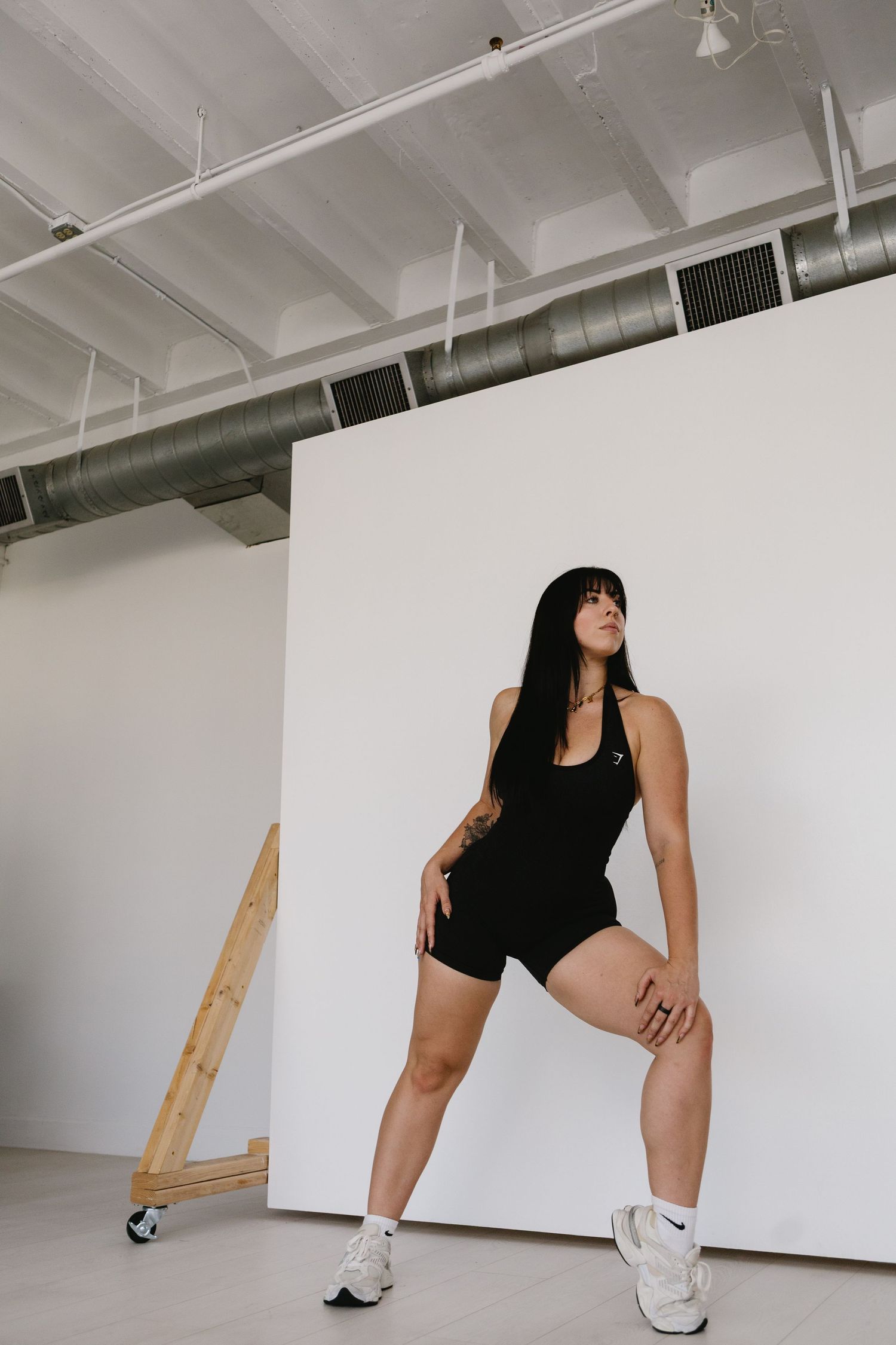 Dark-haired woman in black athletic romper and white sneakers stretching leg in bright studio space.