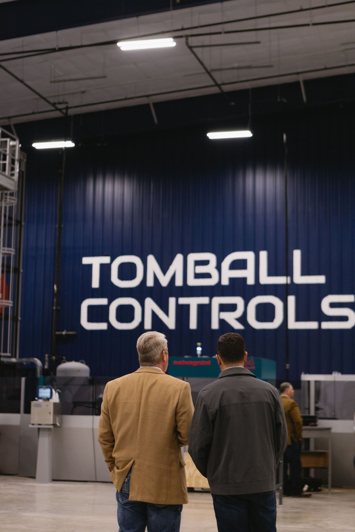 Two people stand in front of a large blue wall with white text reading 'TOMBALL CONTROLS' in an industrial space.