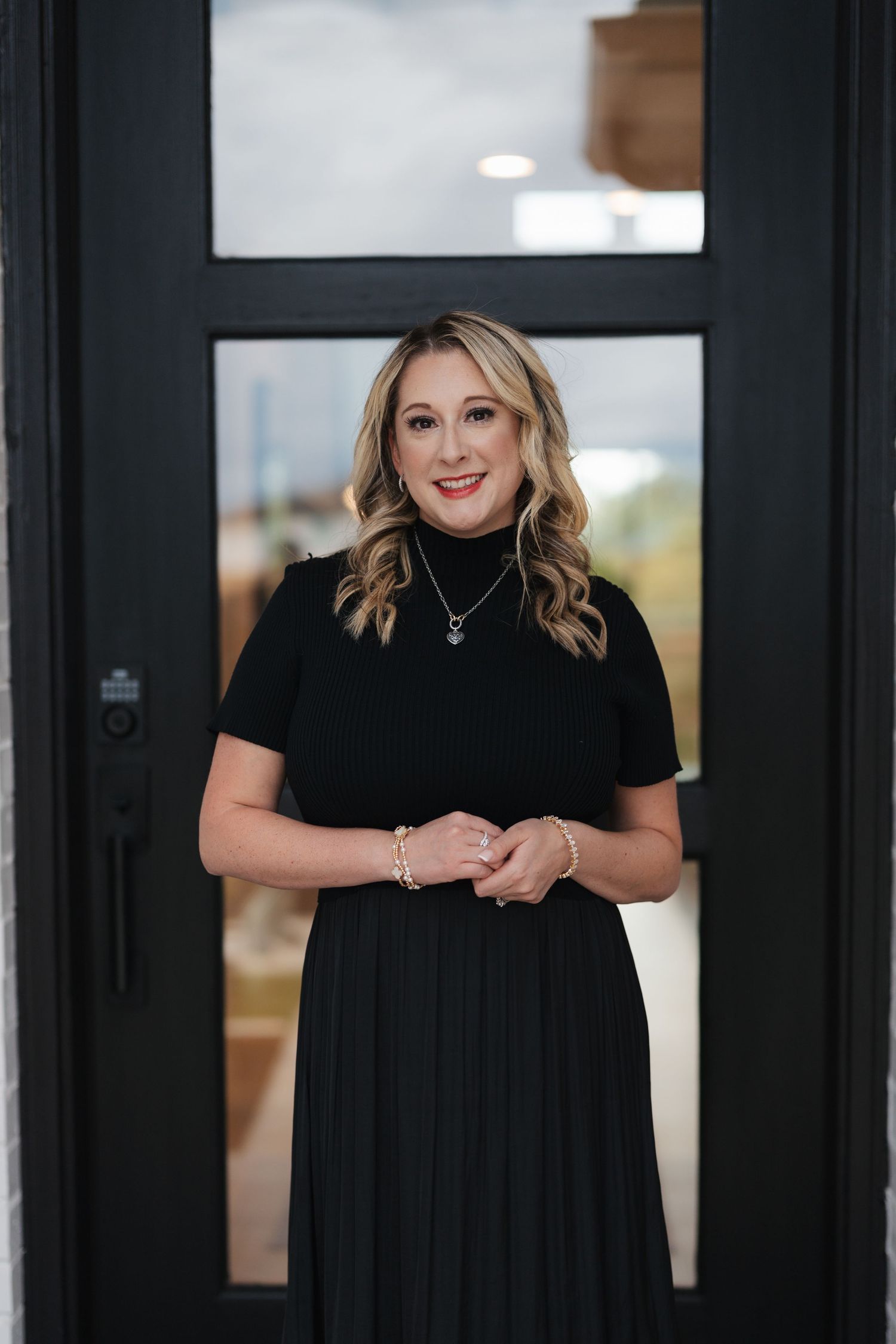 Woman in black dress stands smiling in doorway with pleated skirt and short sleeves.