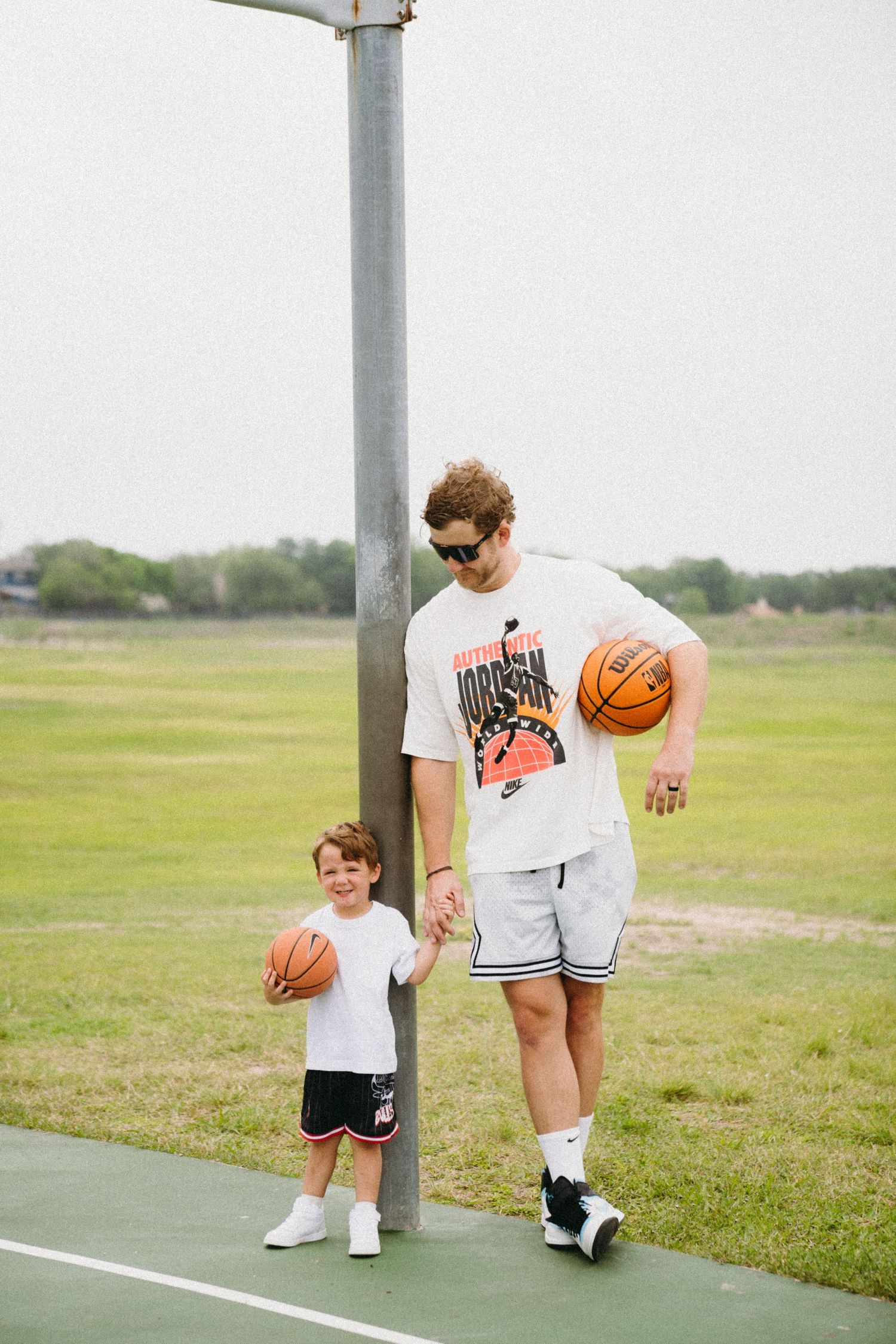 A man and young boy holding basketballs stand together on an outdoor court, both wearing matching white shirts and black shorts.