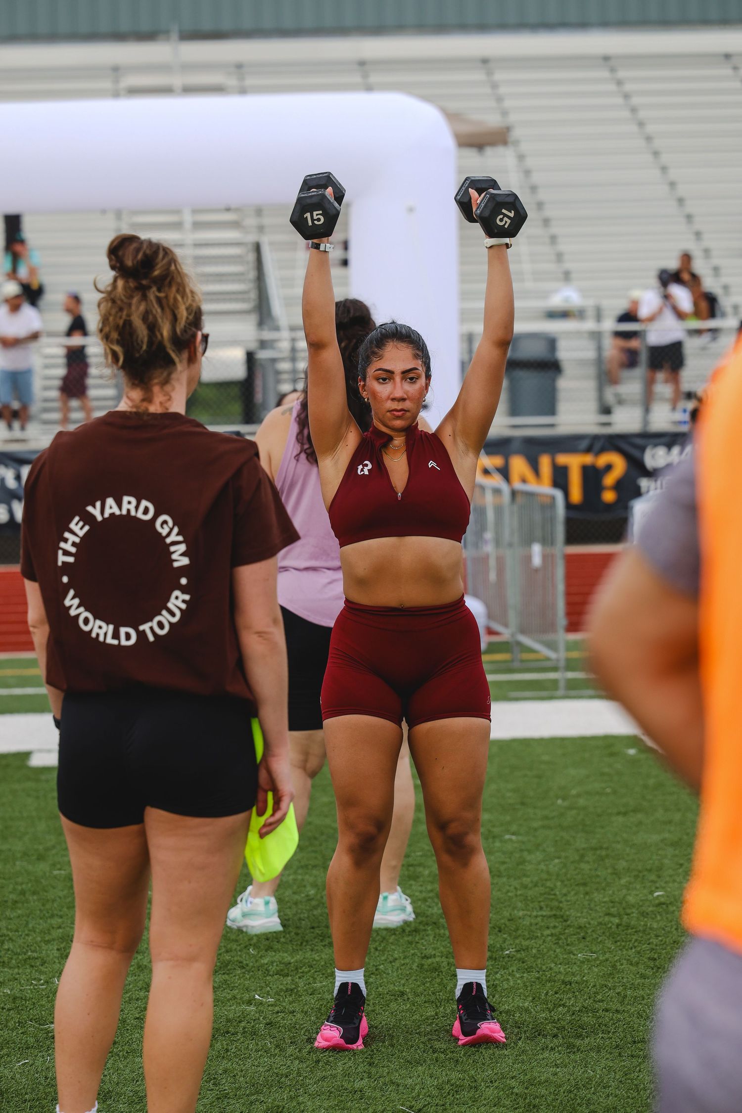Woman lifting dumbbells overhead at The Yard Gym World Tour outdoor fitness event on a sports field.