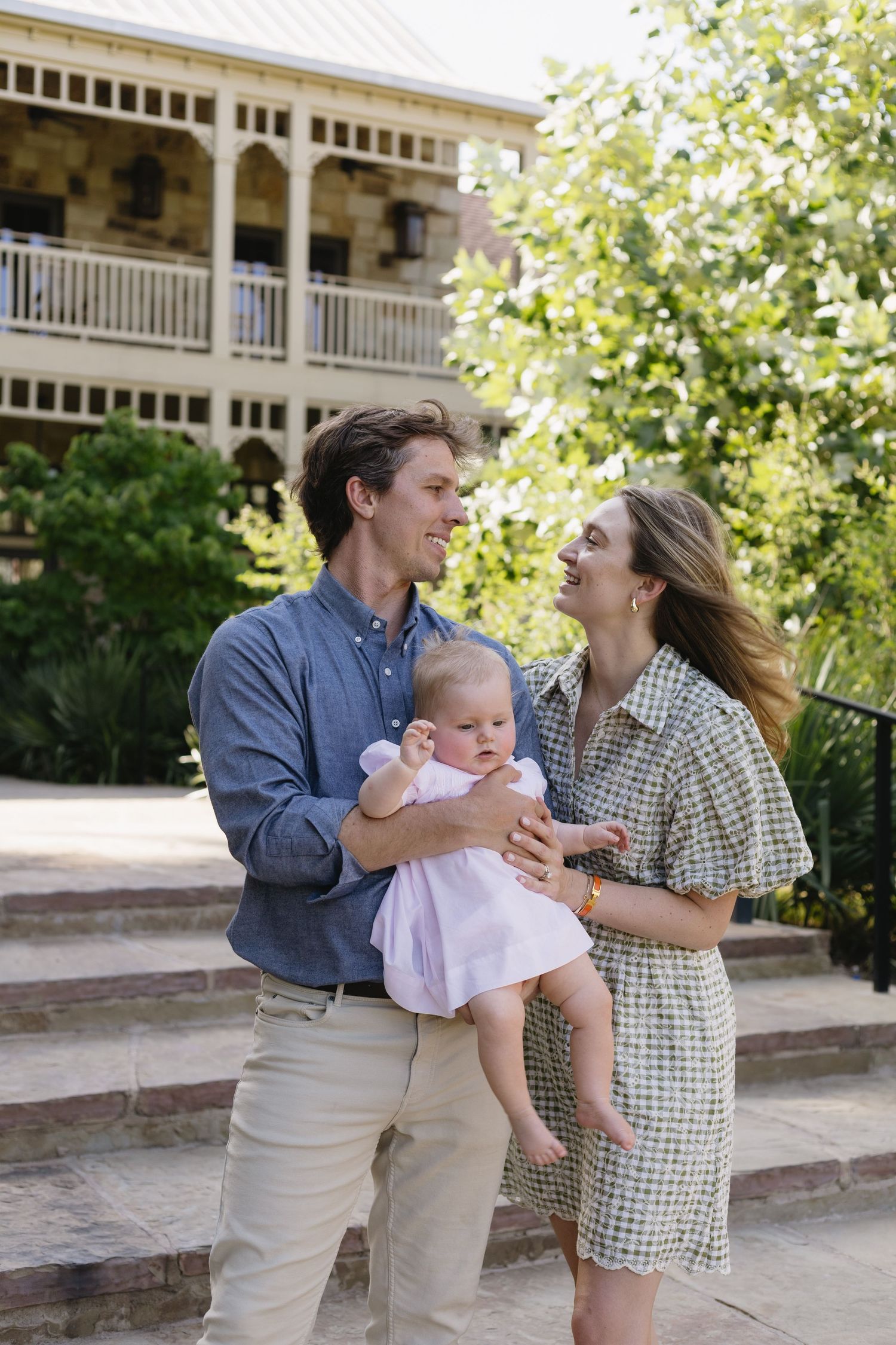 Young couple smiling at each other while holding their baby girl outdoors near a historic building.