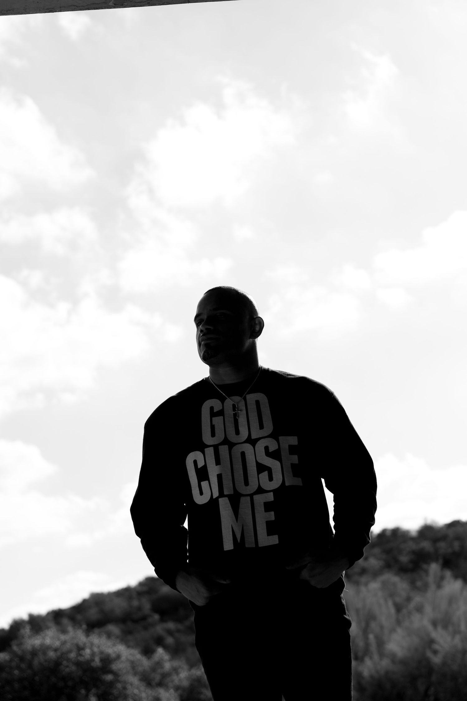 Black and white silhouette of a man wearing a God Chose Me sweatshirt against a cloudy sky.