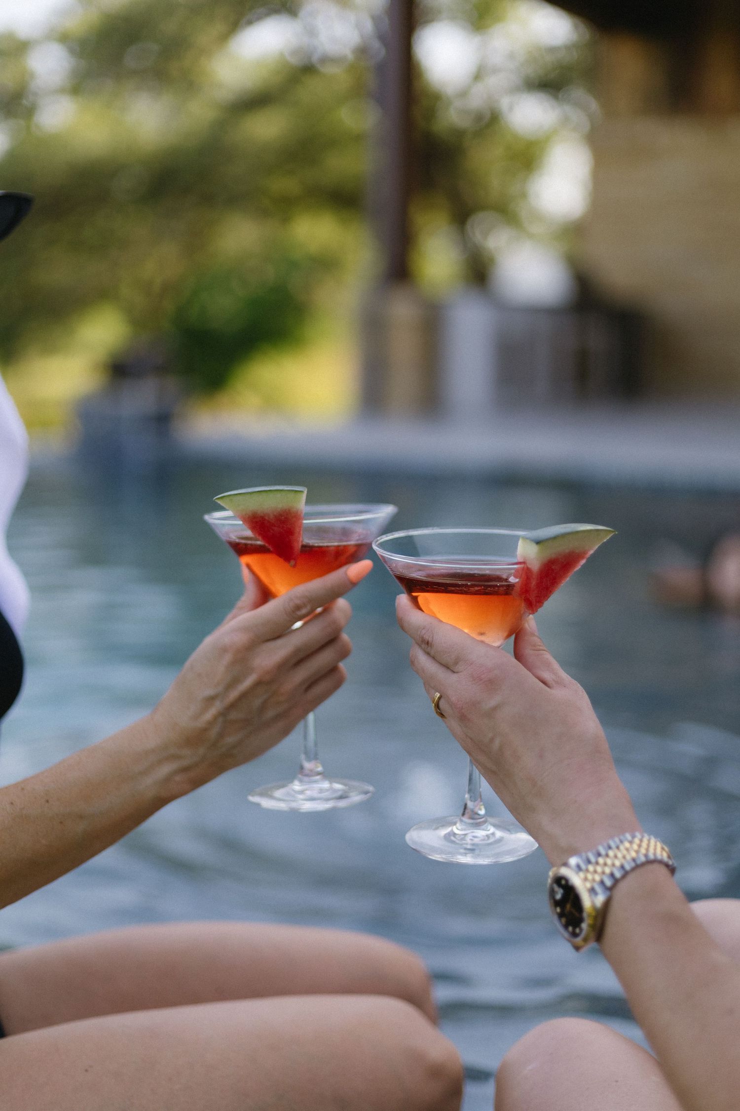 Two hands holding cocktail glasses toasting by a swimming pool.