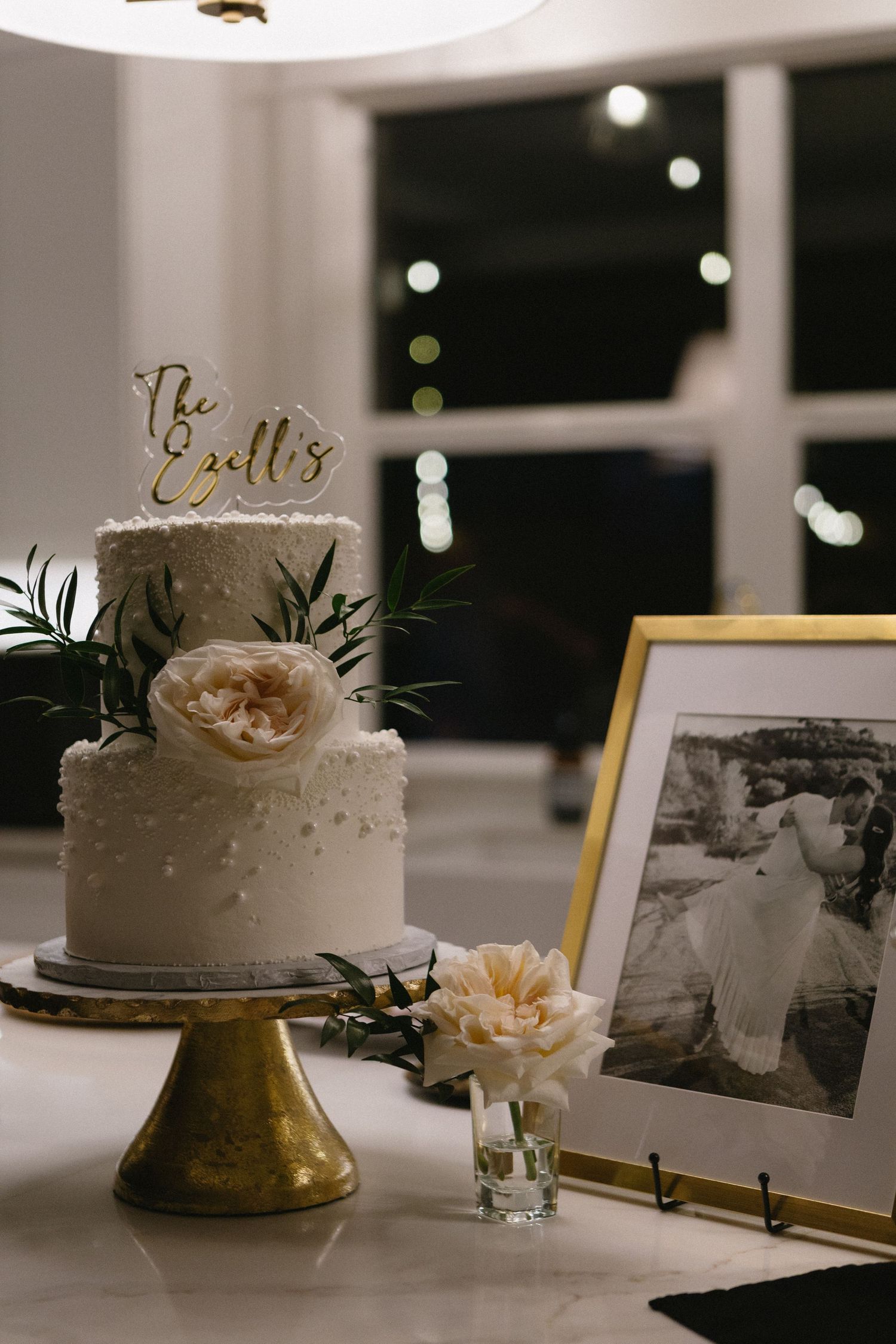 Two-tier white wedding cake with gold topper reading The Ellis, adorned with blush peonies on a gold cake stand.