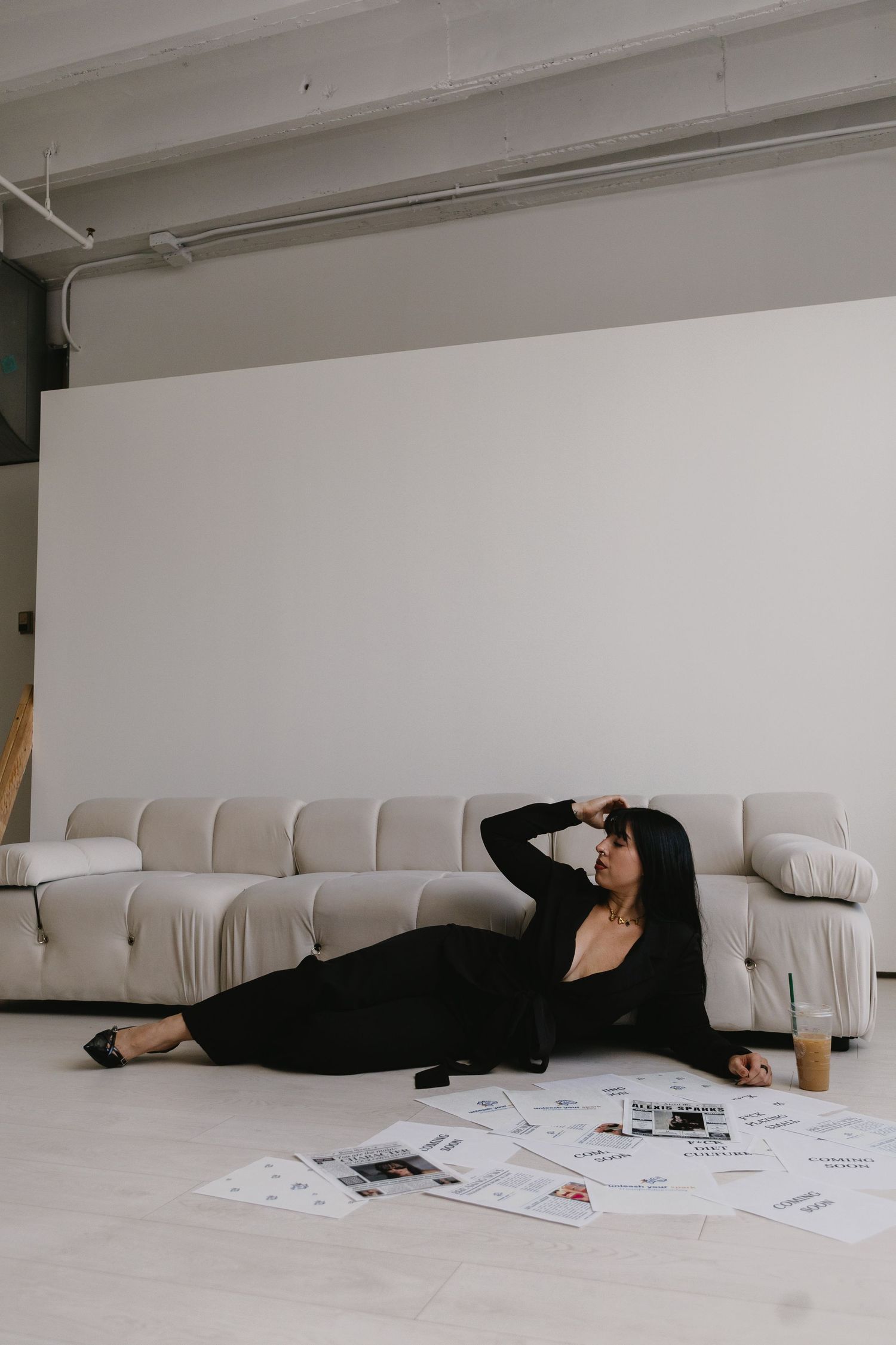 Woman in black outfit reclining on floor near white sofa surrounded by printed photos in modern studio.