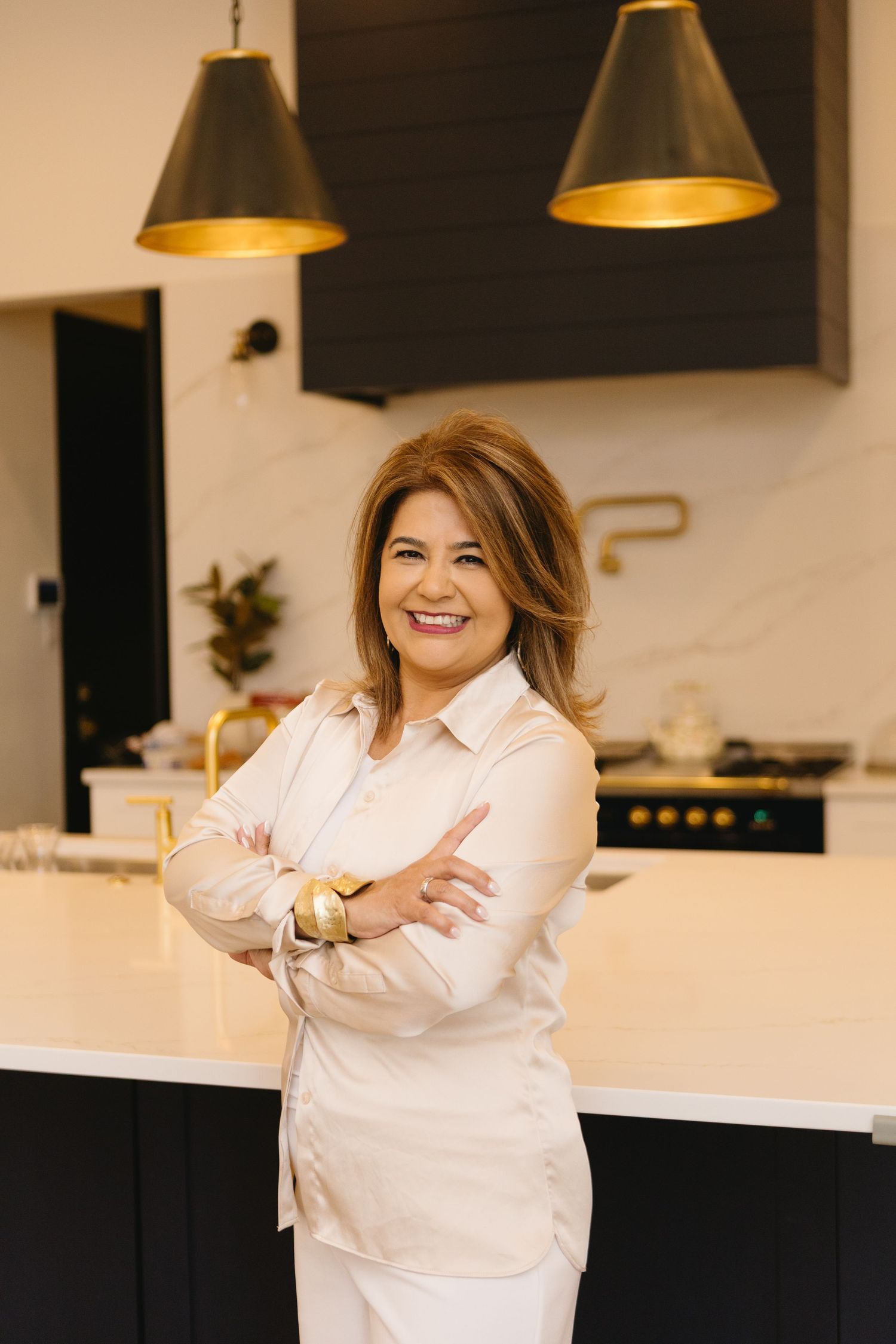 Smiling woman in white blouse with arms crossed standing in a modern kitchen with black and gold pendant lights.