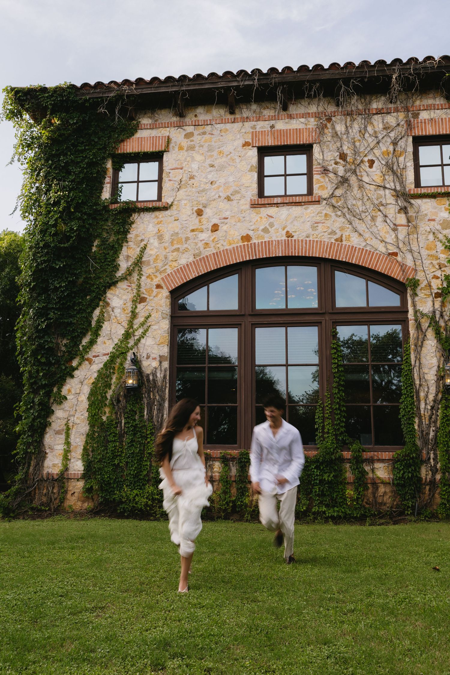 Couple in white outfits running on green lawn before ivy-covered stone villa with arched windows.