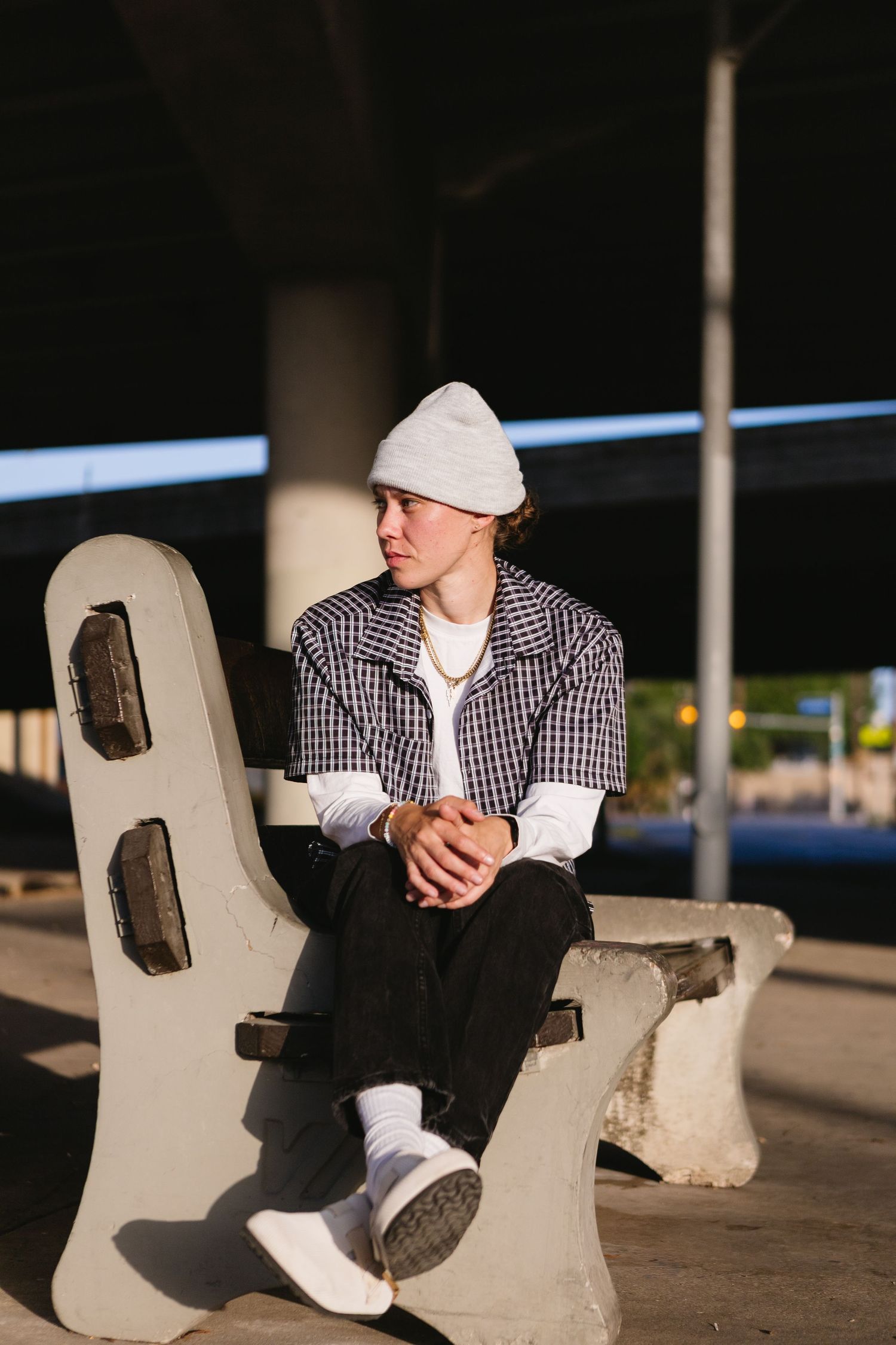 Young man in plaid shirt and white beanie sits on concrete bench under urban overpass, gazing thoughtfully to the side.