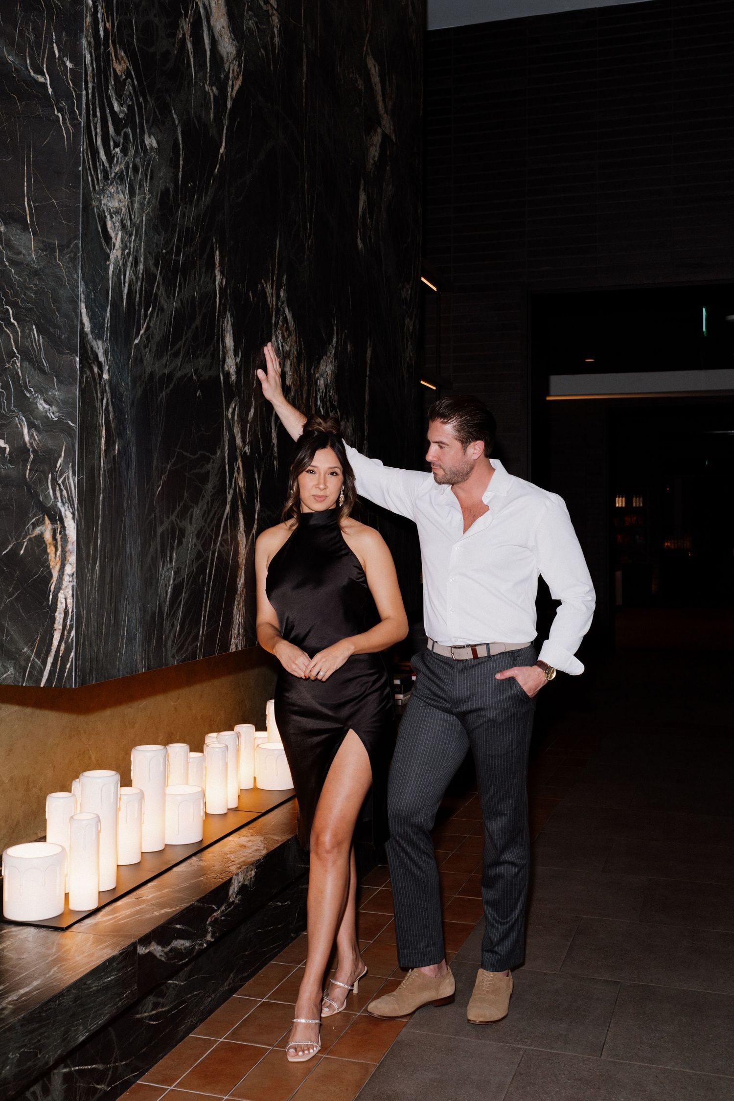 Elegant couple in formal wear posing near candlelit wall in upscale venue.