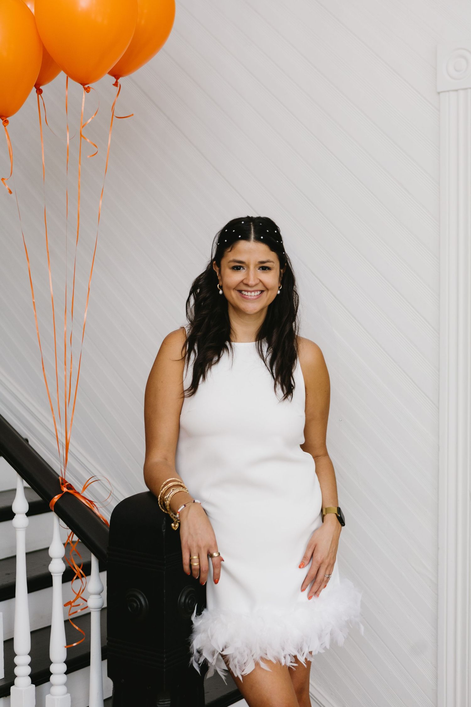 Woman in white feathered dress poses on stairs with orange balloons for celebratory photoshoot.