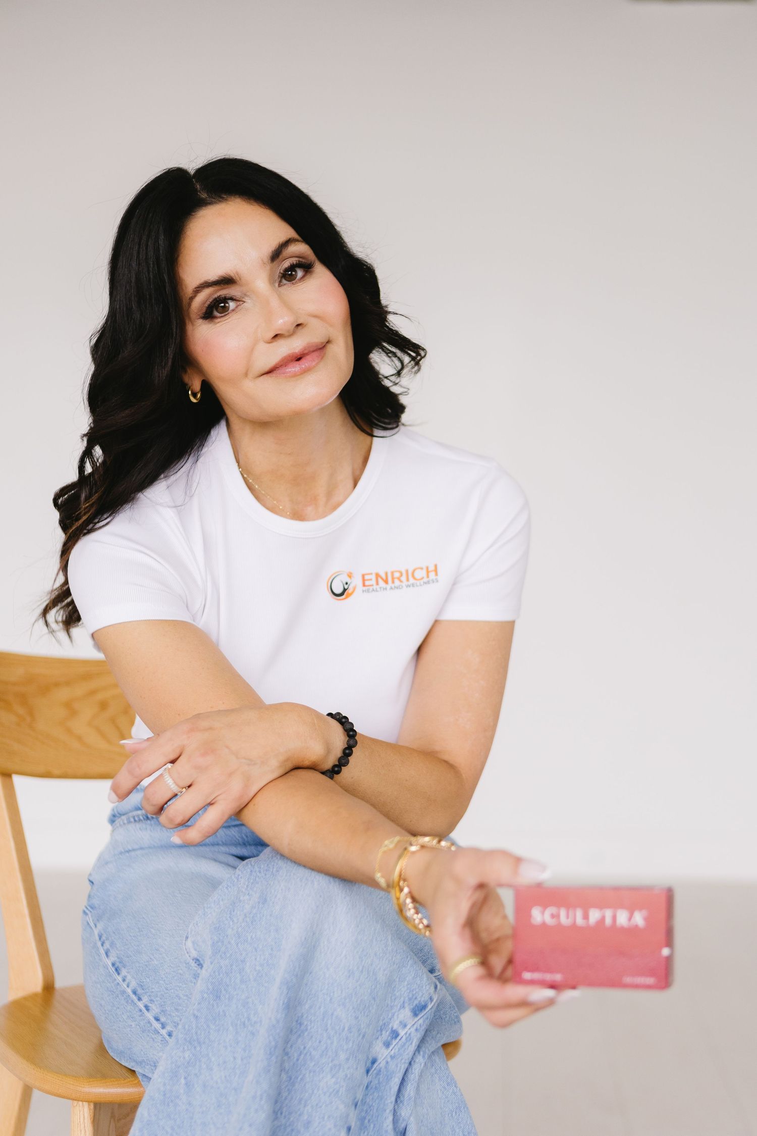 Woman in white Enrich t-shirt sitting on wooden chair holding Sculptra product, smiling confidently.