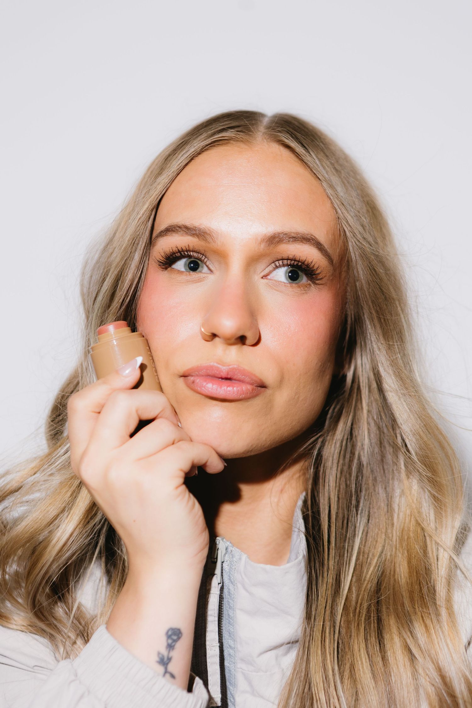 Young blonde woman holding a makeup foundation bottle against her cheek, looking up thoughtfully against white background.