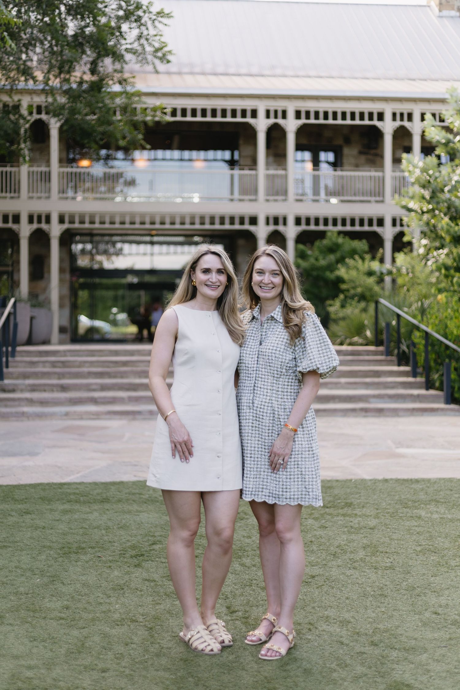 Two young women in summer dresses smile together outdoors in front of a historic two-story building.