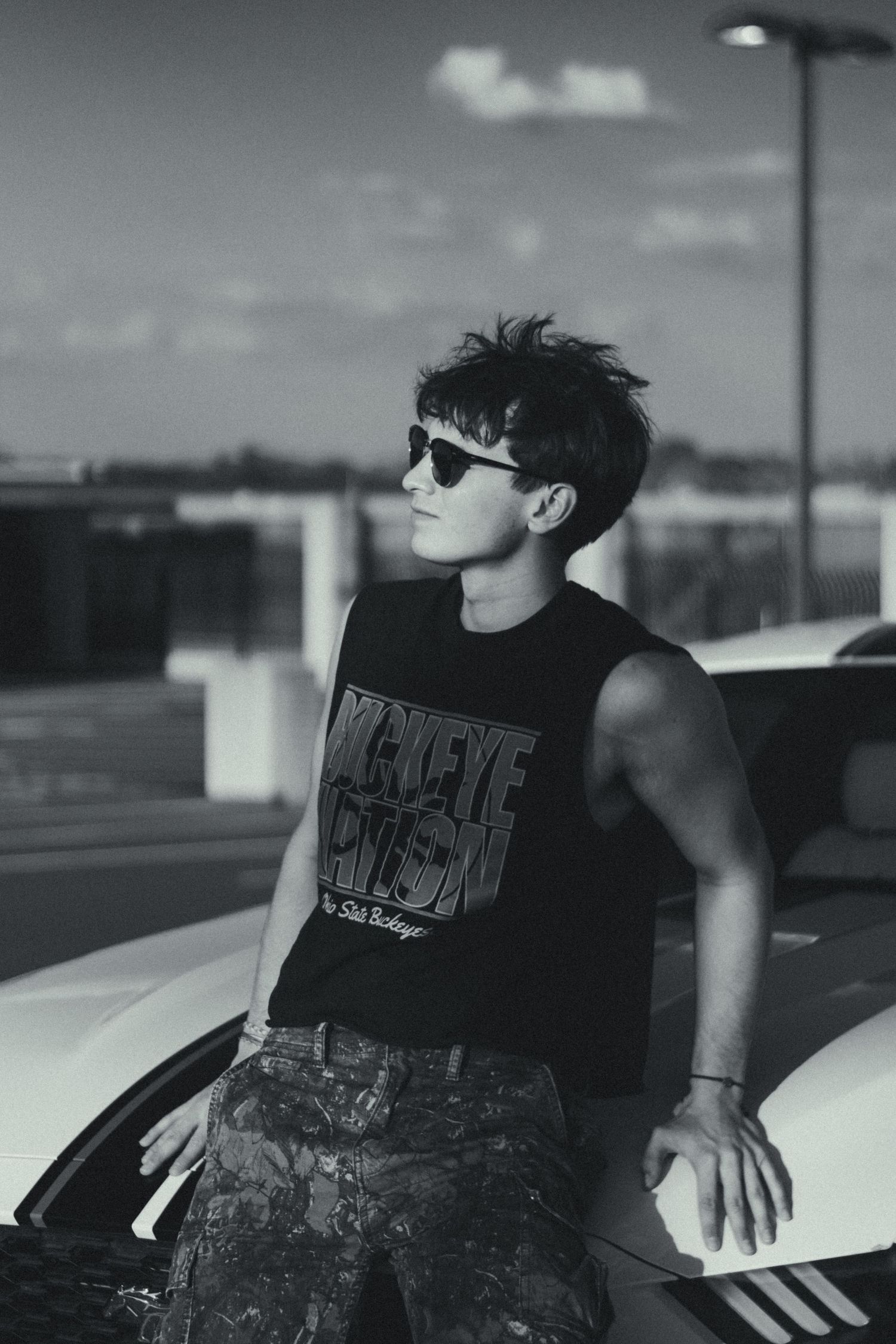 A black and white photo of a man lean on the hood of his car.