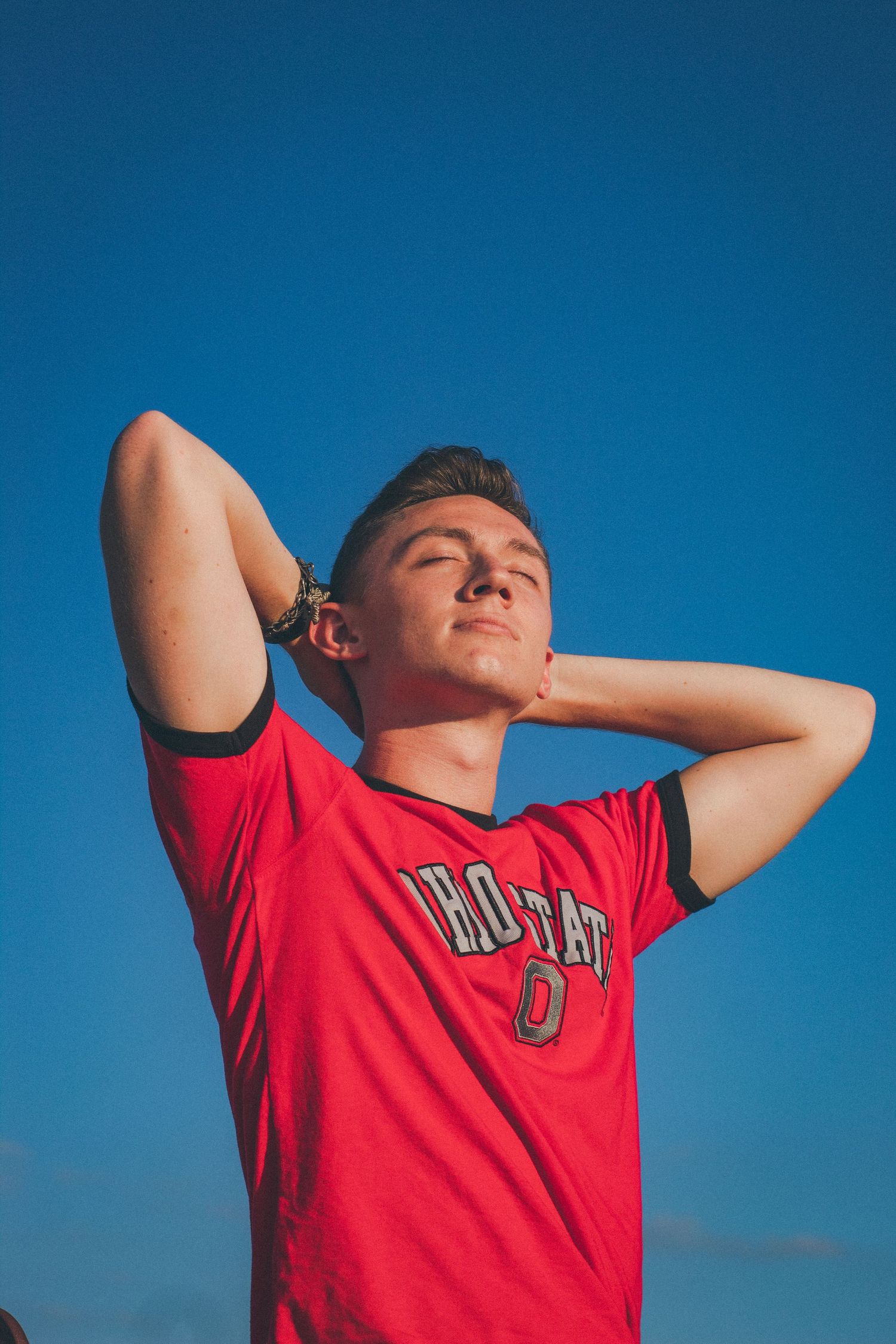 A man holding his hands behind his head in front of a blue sky.