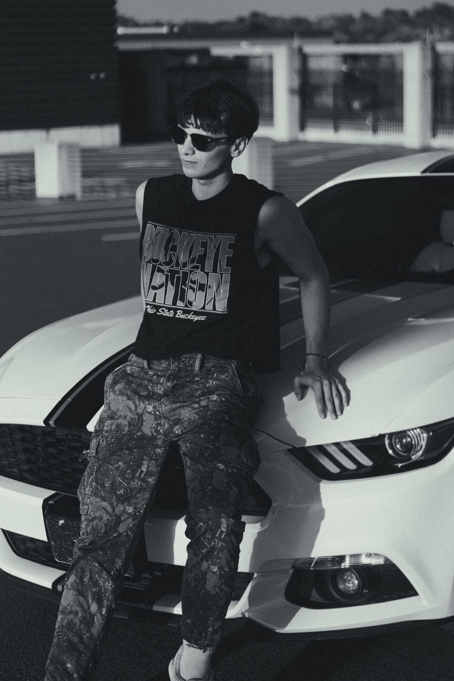 A black and white photo of a man leaning on the hood of his car on the top of a parking garage.