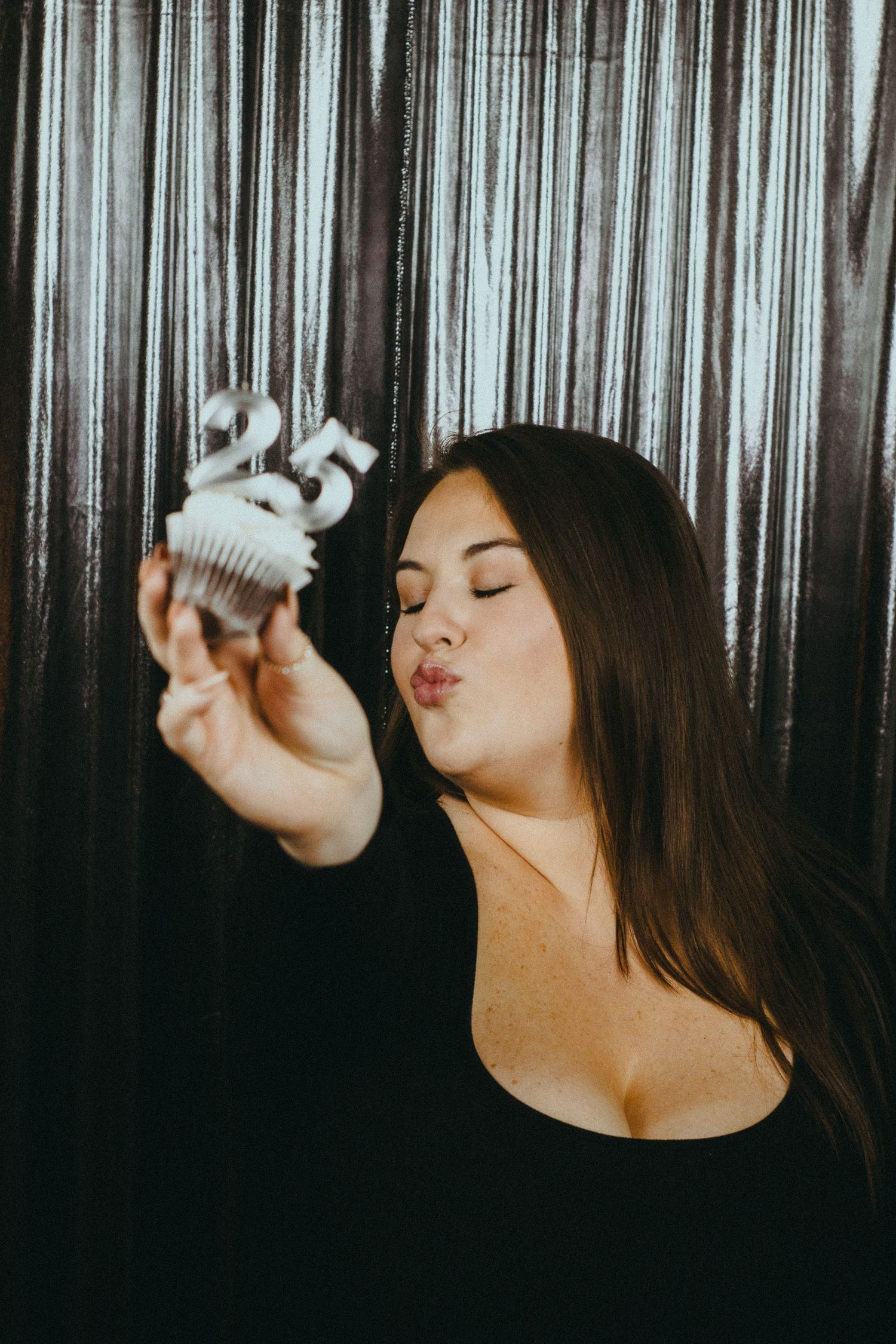 A photo of a woman standing in front of a silver, shimmering backdrop and holding a cupcake to the camera.