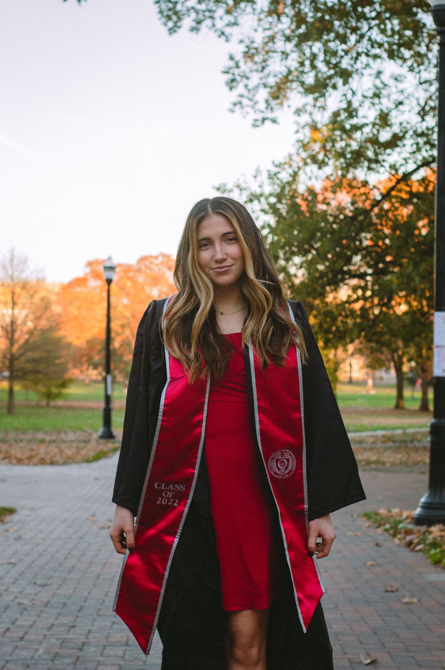 A woman in a graduate gown walking towards the camera in front of trees.