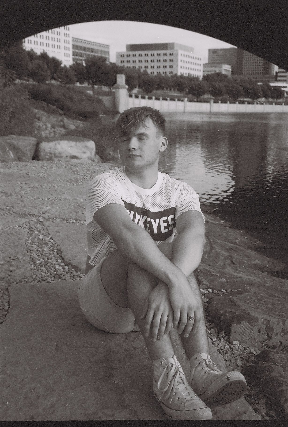 A black and white photo of a young man sitting near a river. A bridge is behind him.