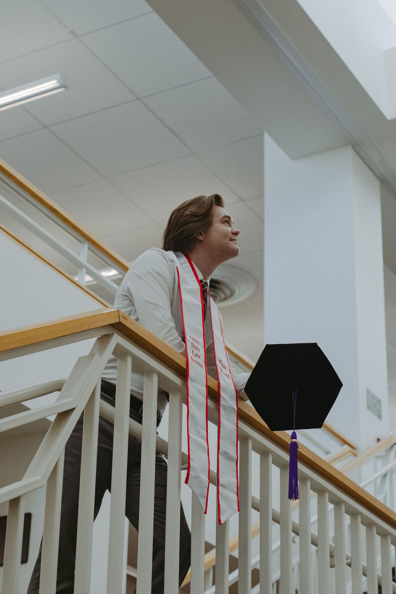 A man with a graduation cap leans on a staircase railing.