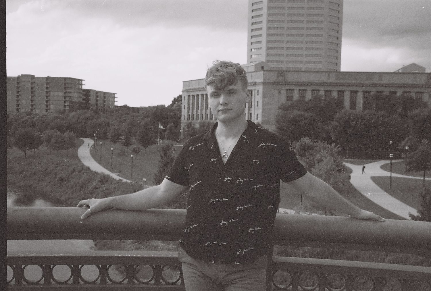 A black and white photo of a young man standing in front of a railing, a cityscape behind him.