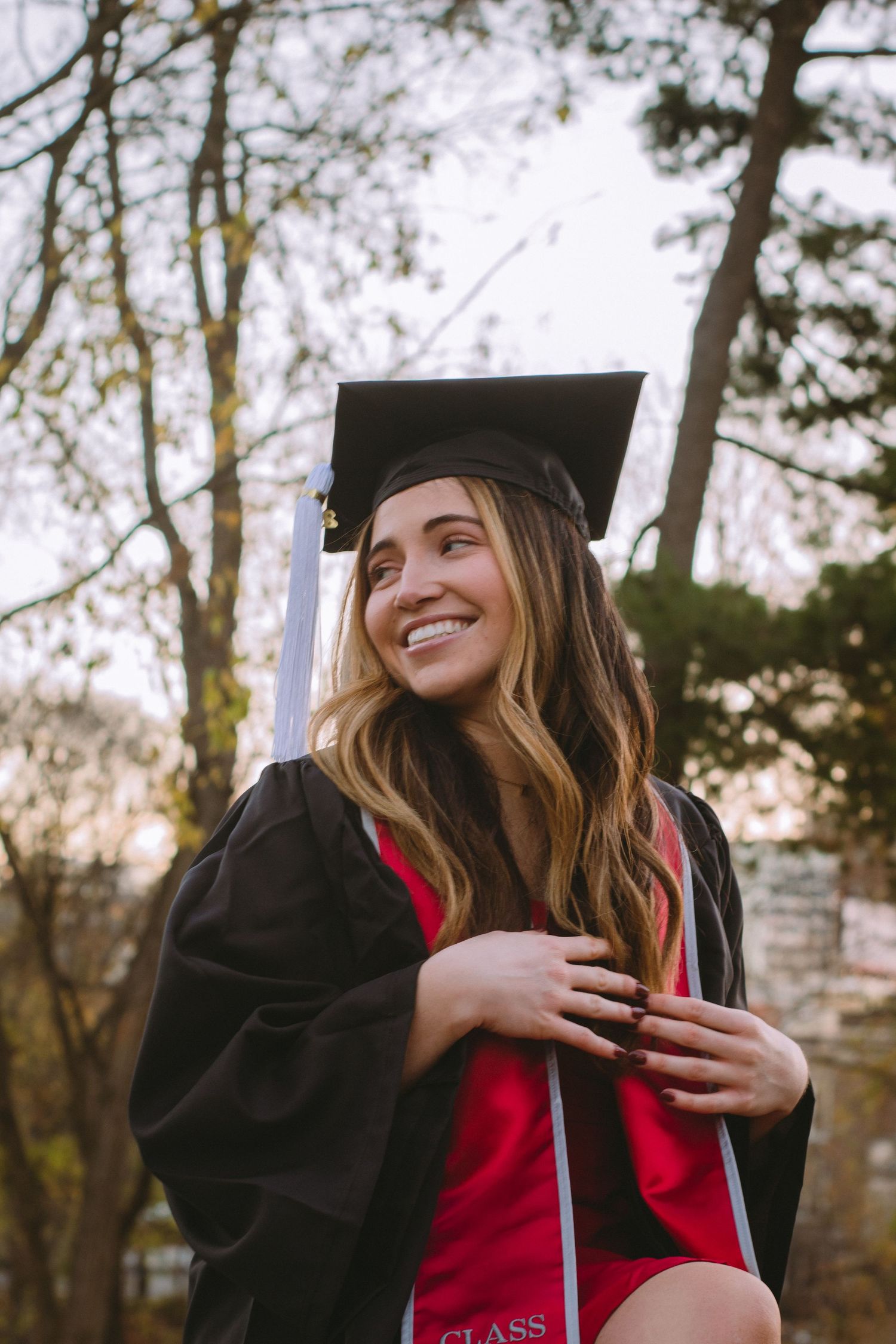 A woman wearing a graduation cap and gown in front of a trees and a blue sky.