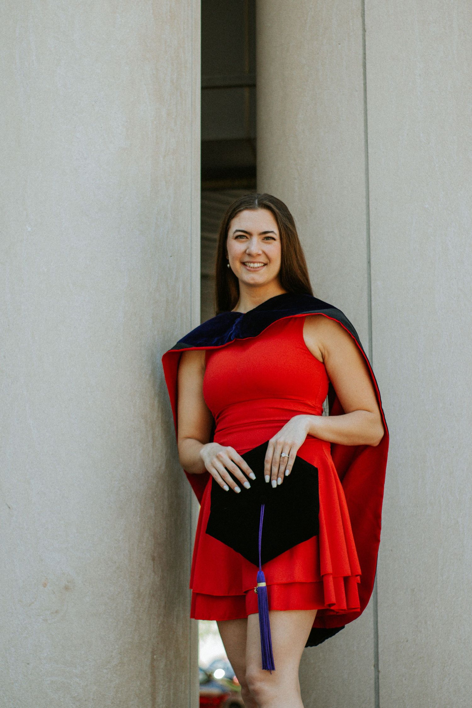 A woman in a red dress standing in front of a concrete pillar holding a graduation cap.